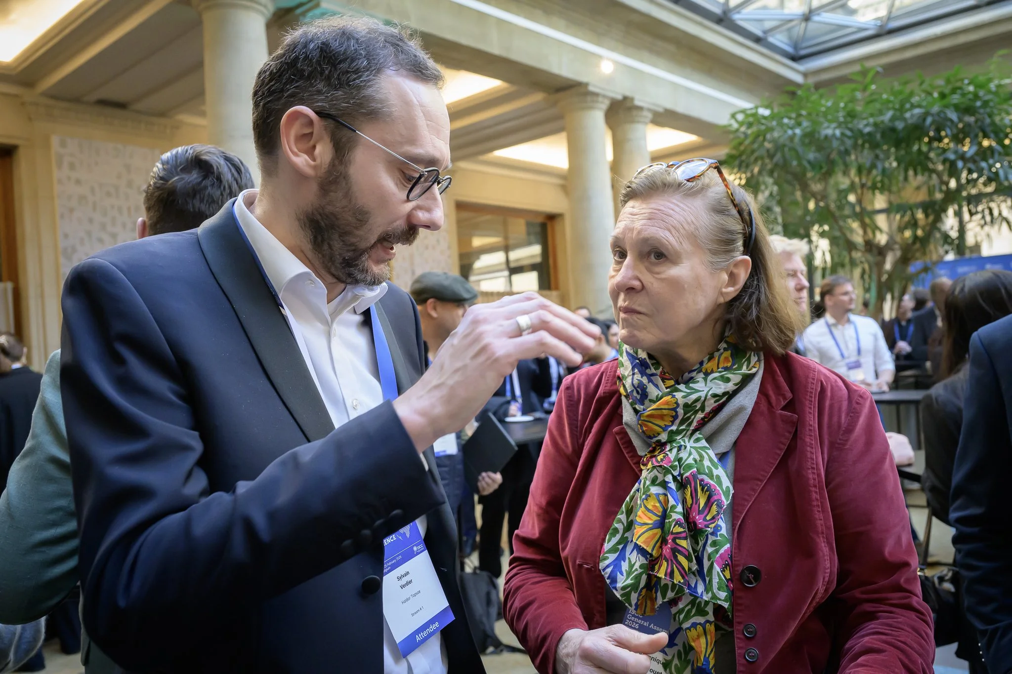 A man with glasses gestures while speaking to a woman in a red jacket and patterned scarf.