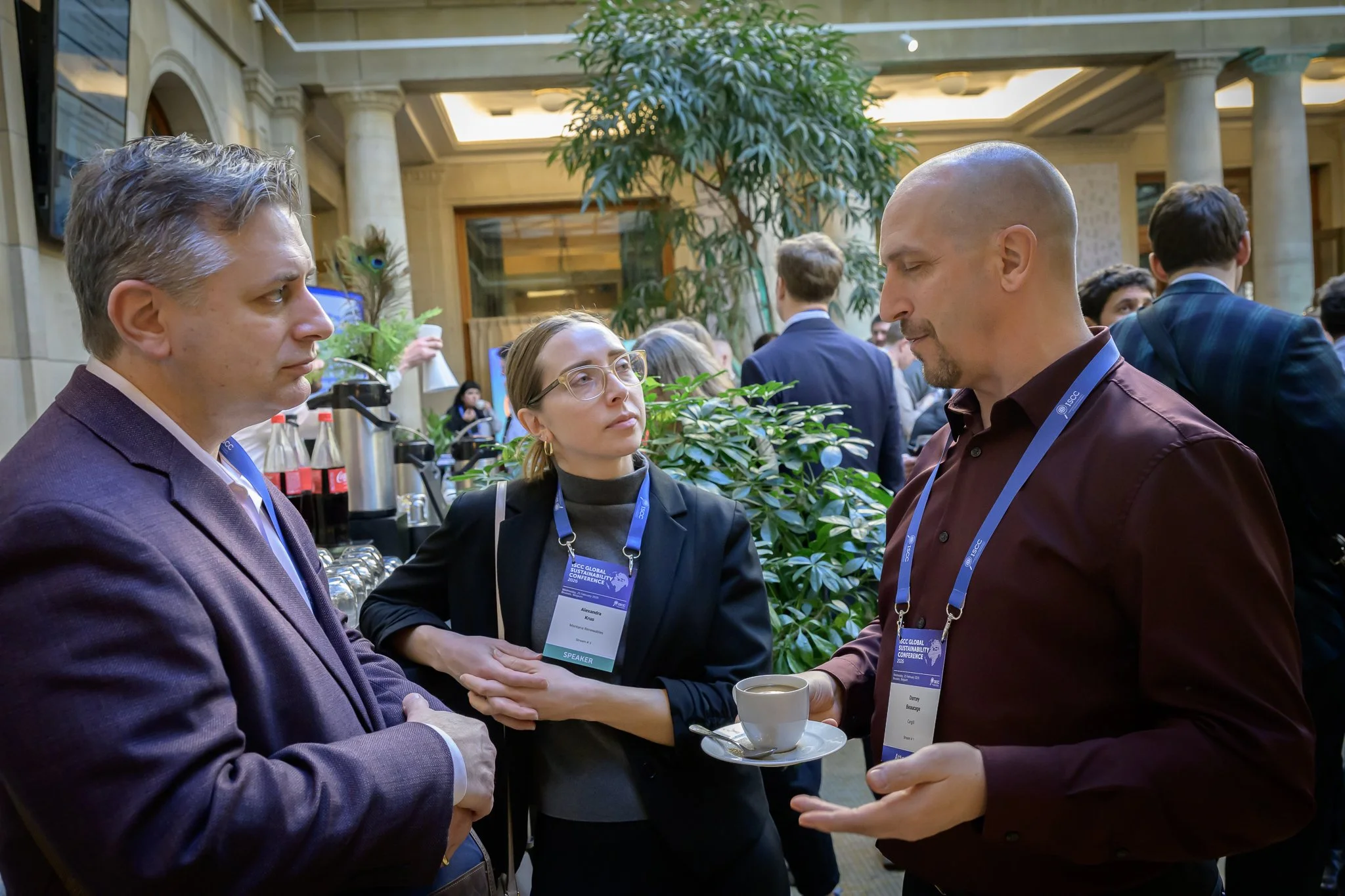 Three participants talk in the conference lobby, one holding a cup and saucer.