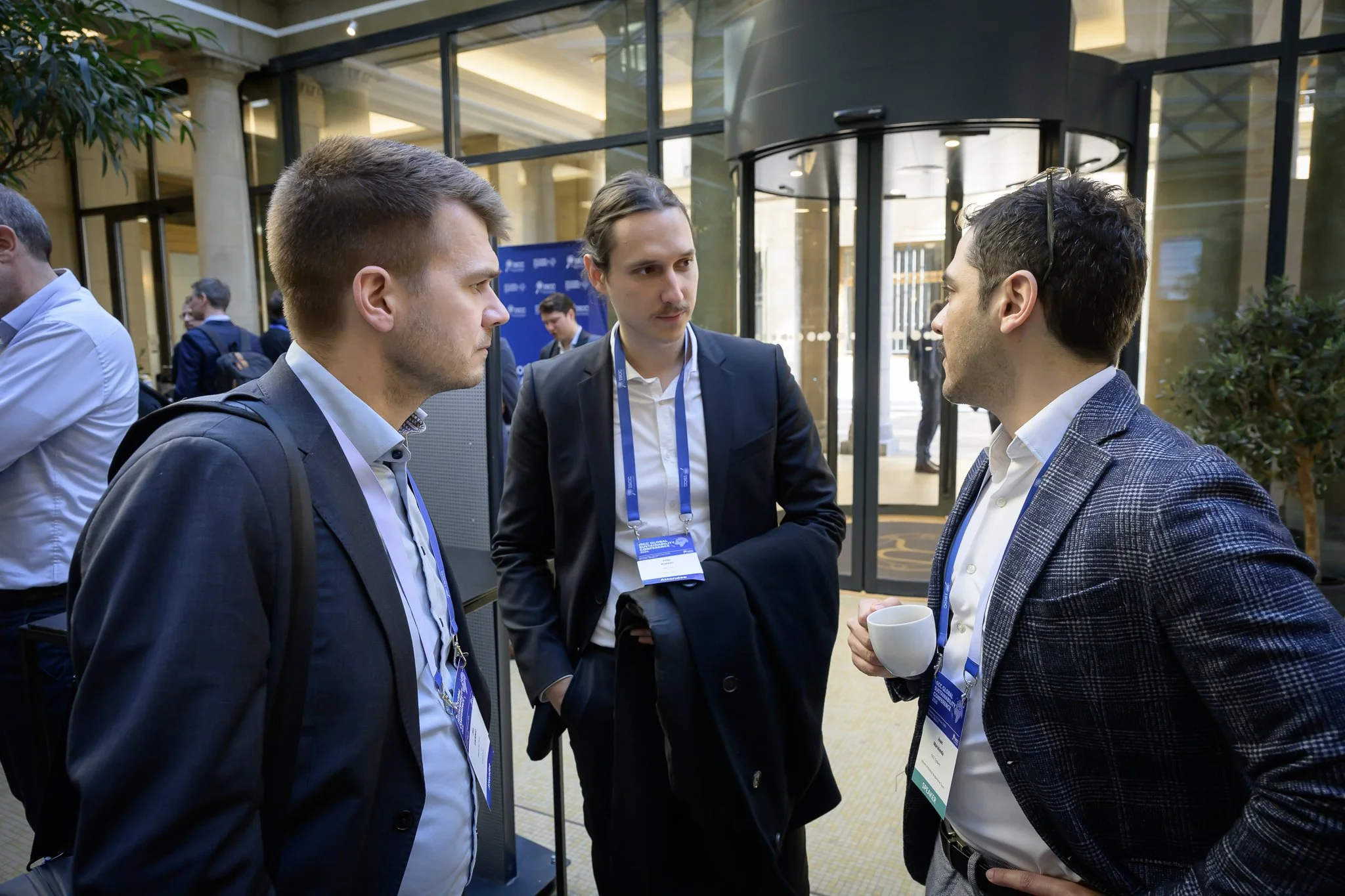 Three men wearing conference lanyards converse in the bright lobby.