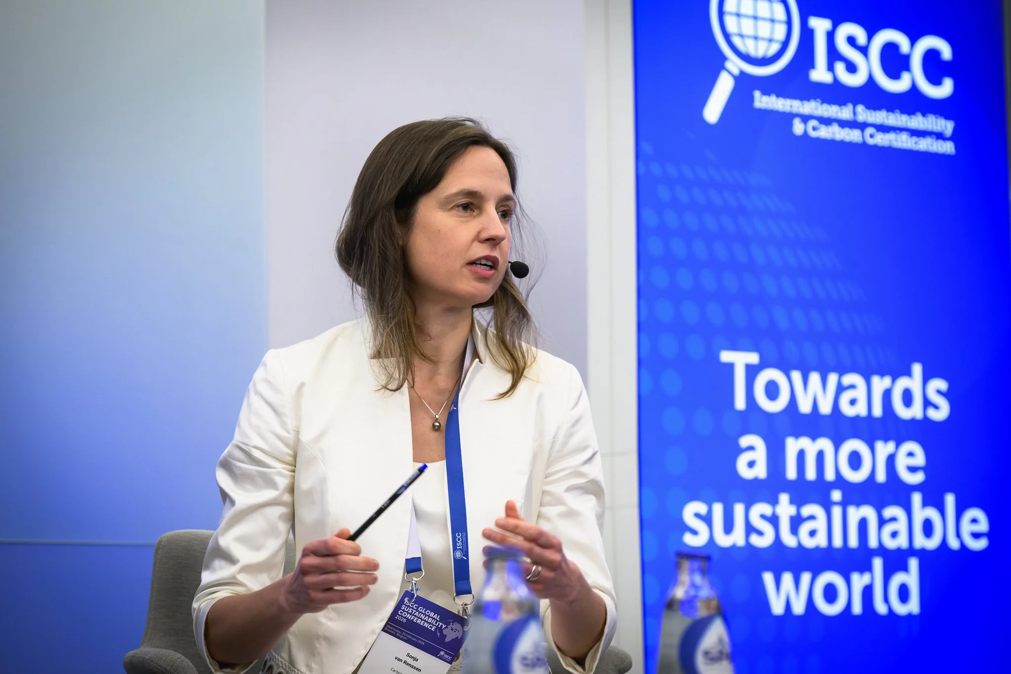 A seated woman in a white jacket gestures beside an ISCC banner.