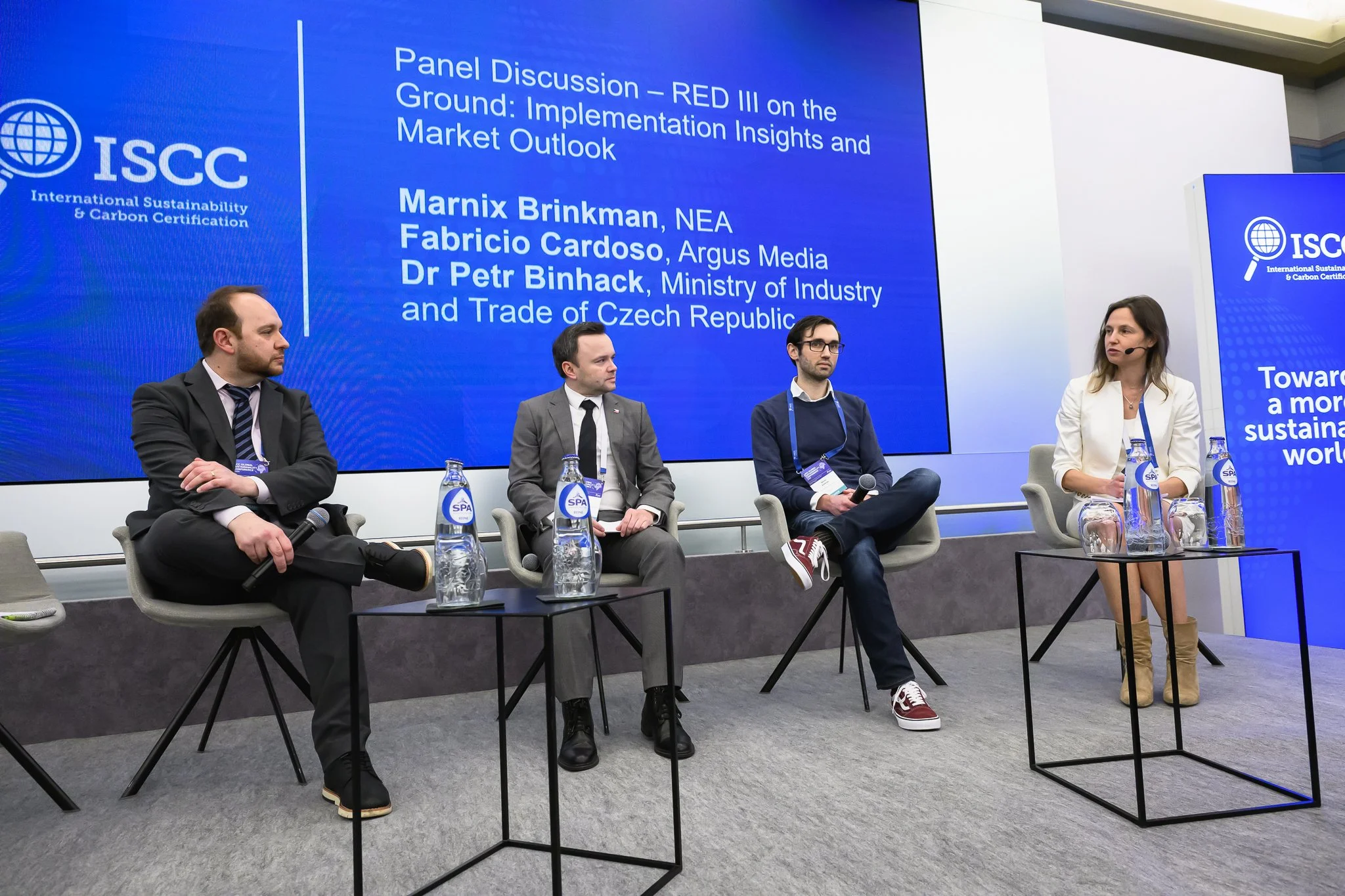 Three men and a woman sit on stage for a panel discussion beneath a blue presentation screen.