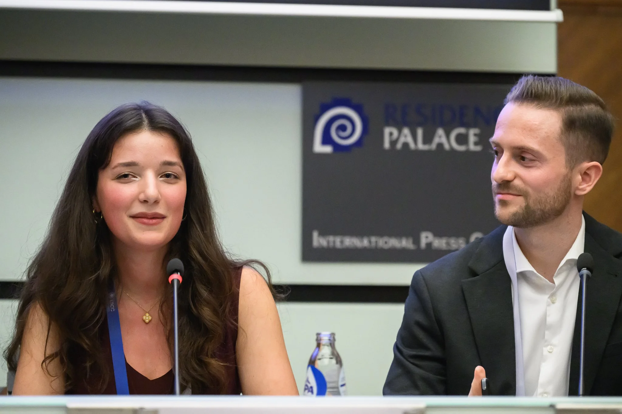 A woman and a man sit side by side at a conference panel table.
