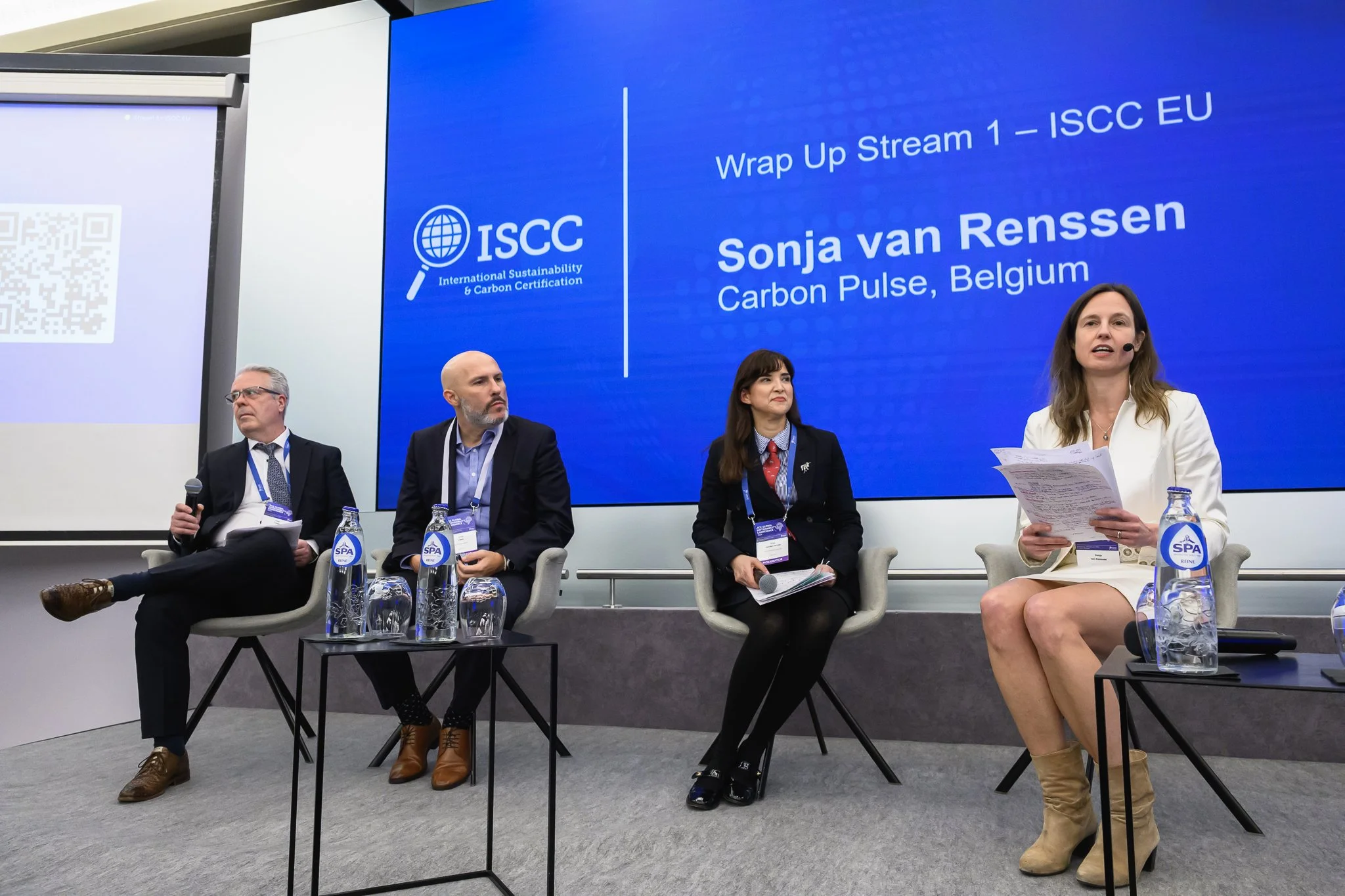 Four panel participants sit beneath a wrap-up presentation slide, with the moderator holding notes.