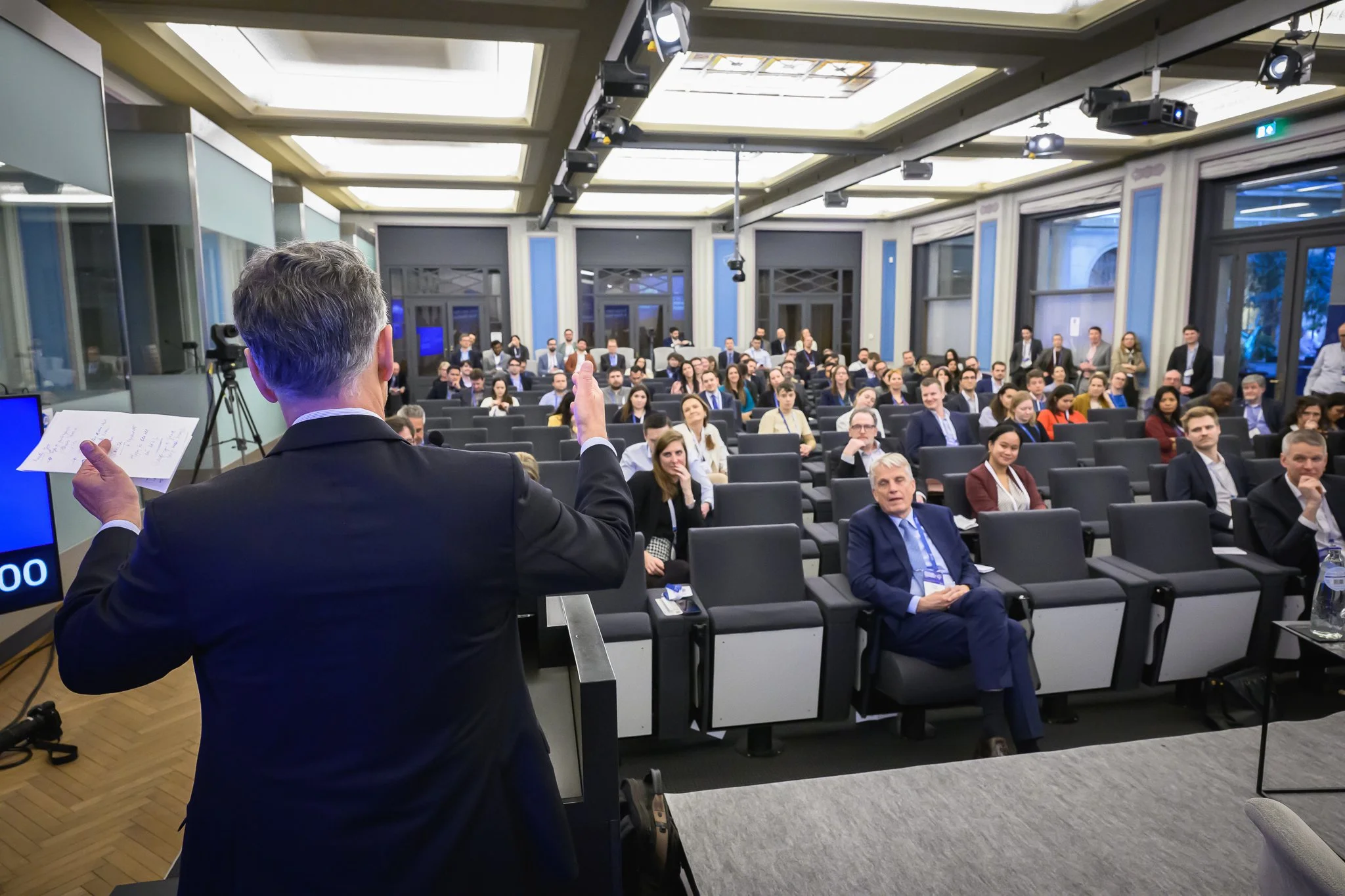 A speaker seen from behind raises one hand towards the audience.