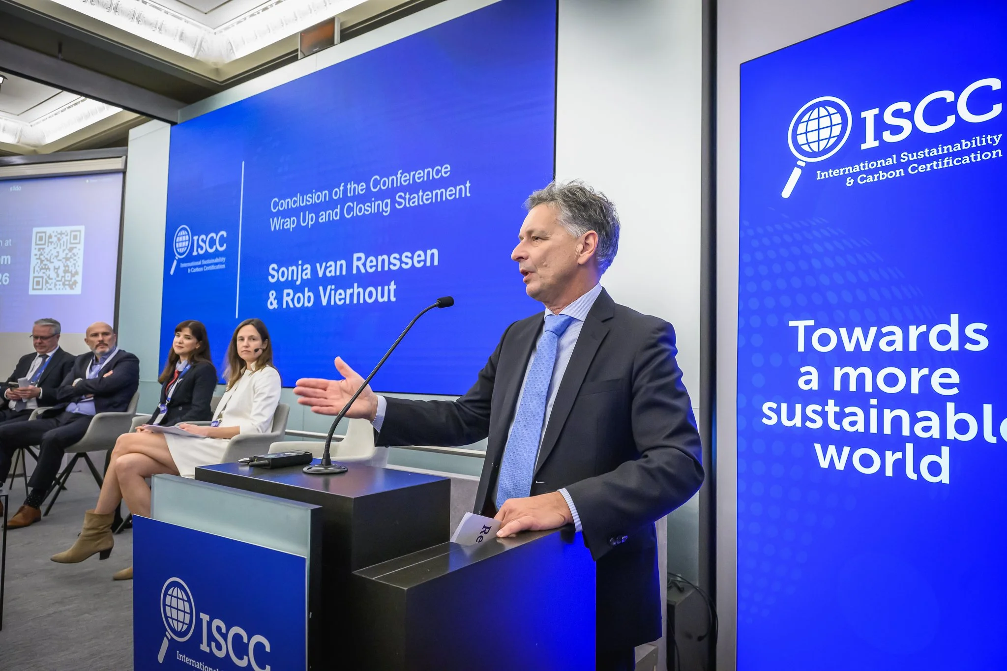 A man gestures at the lectern while panel participants sit behind him.
