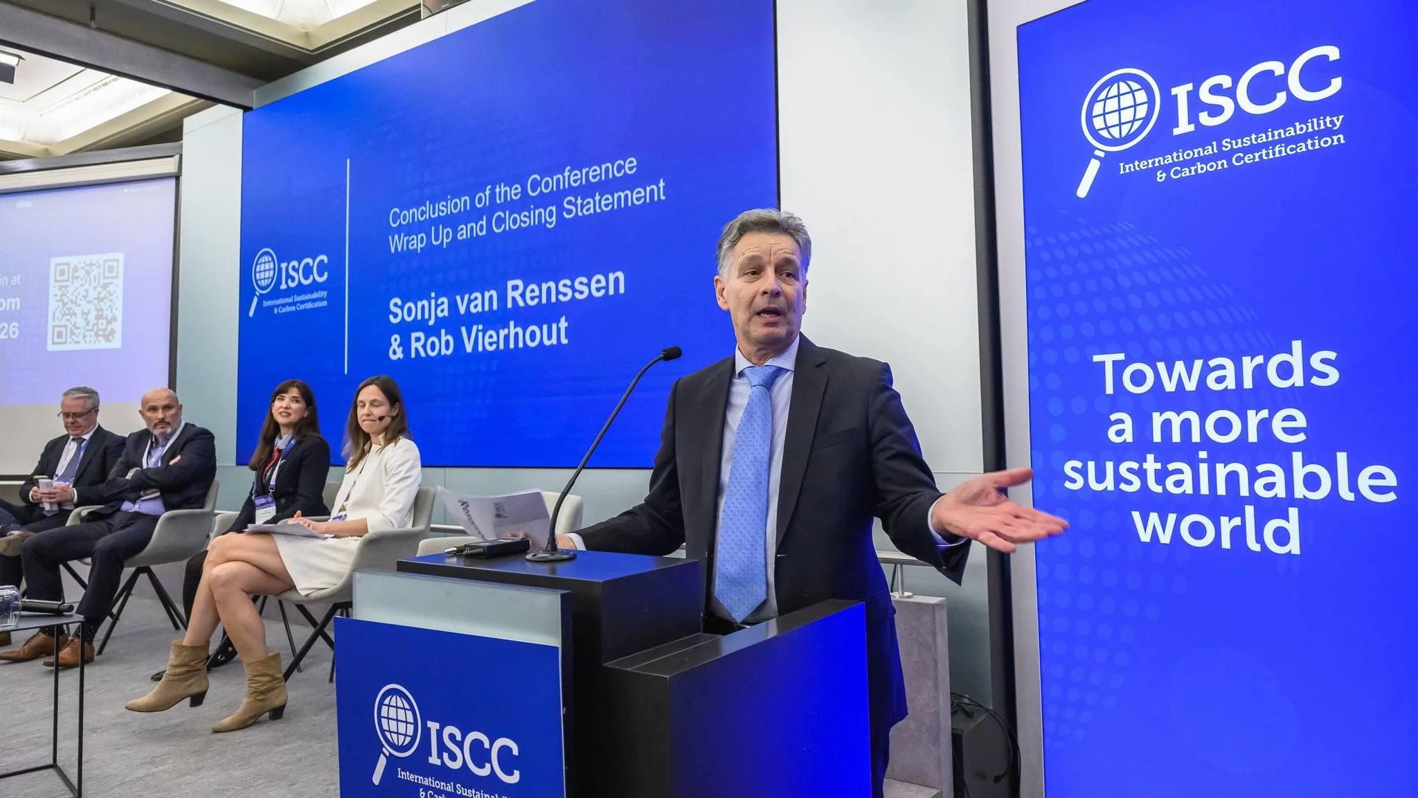 A man at the lectern opens both arms while addressing the audience.