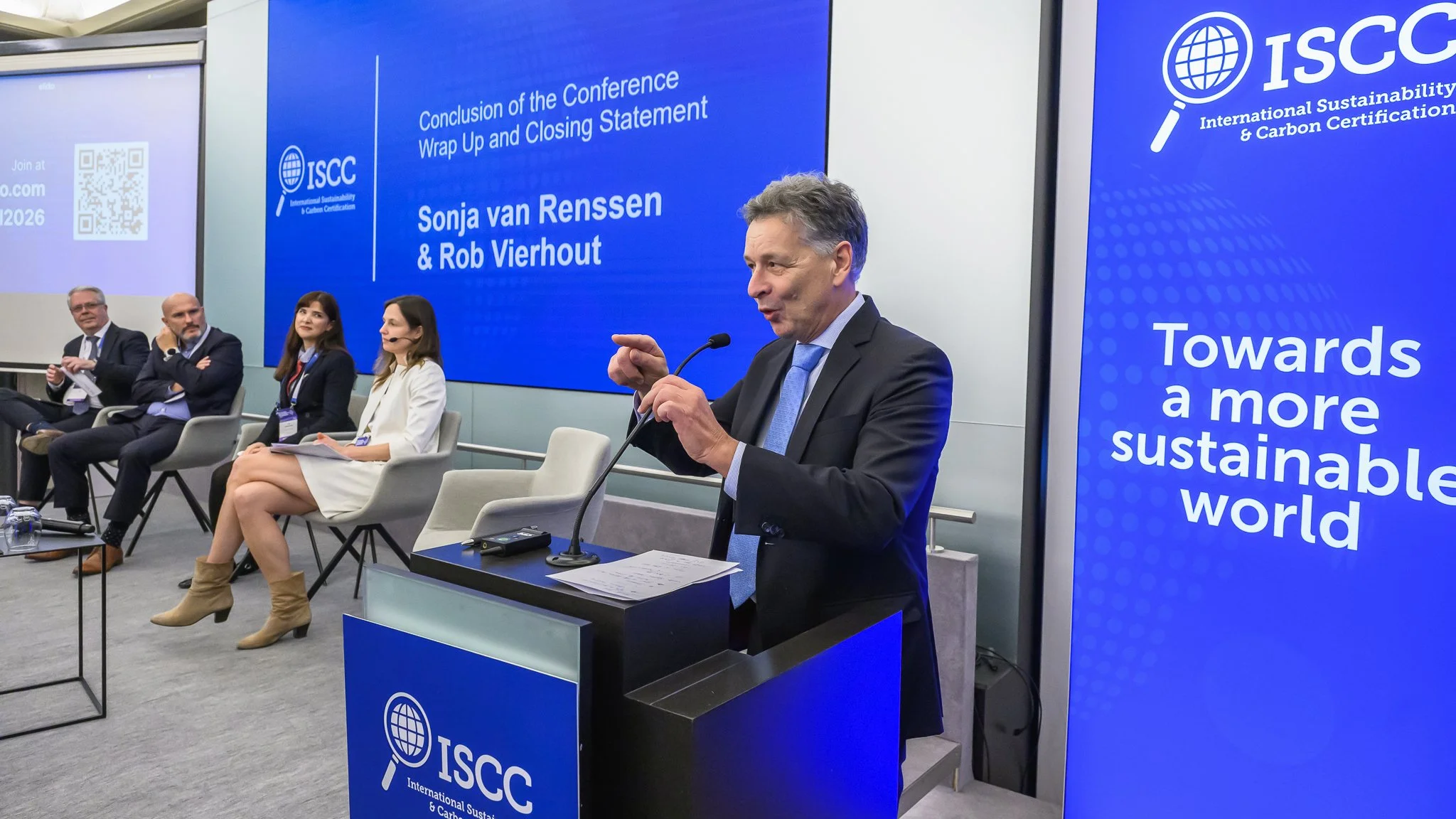 A speaker gestures at the lectern beside a blue ISCC banner during the closing session.