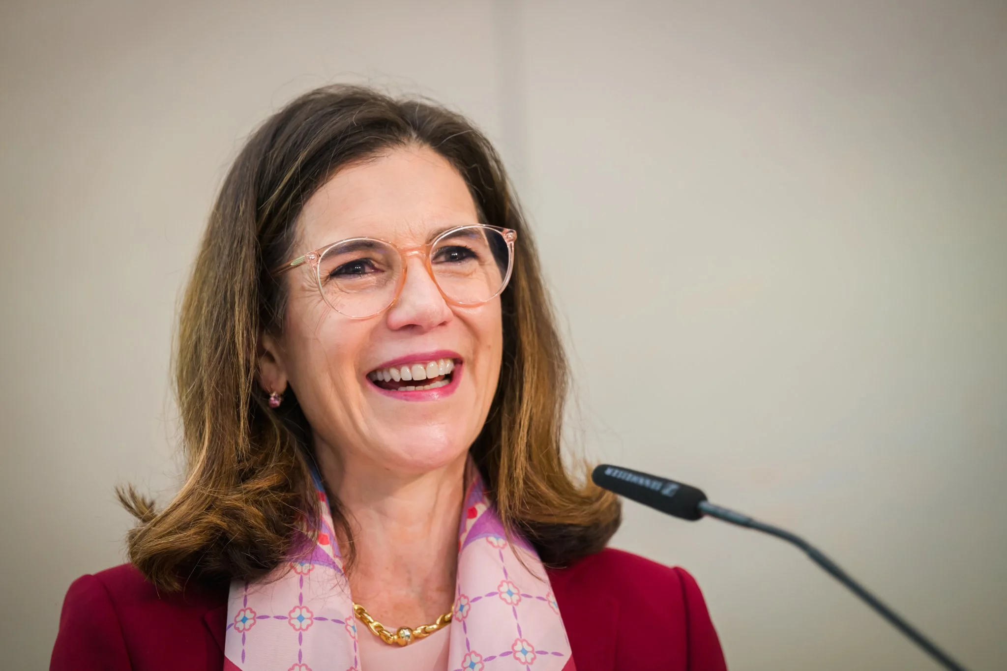 Portrait photo d'une femme souriante portant des lunettes et un foulard rose, tenant un discours devant un micro. Photo réalisée pendant une conférence à Bruxelles.