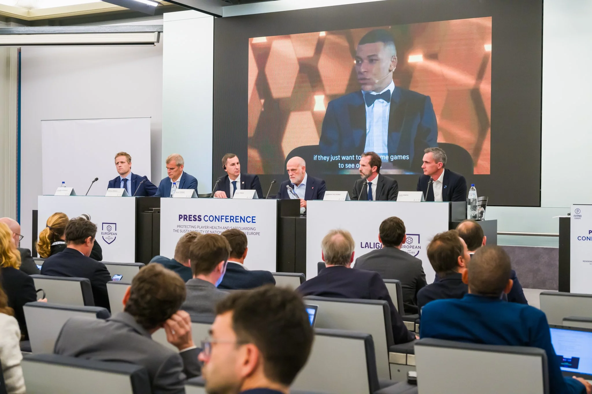 Photo pendant une conférence de presse avec un panel d'hommes en costumes, un grand écran affichant un homme en smoking, et des participants assis. Texte sur le pupitre concernant la santé des joueurs et la durabilité des clubs nationaux en Europe.
