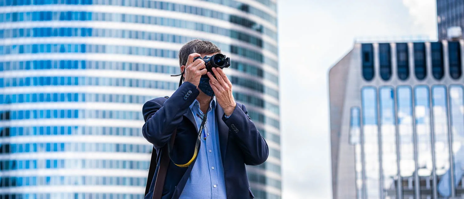 Un homme avec un costume, portant un sac à dos, prend une photo dans un environnement urbain avec des bâtiments modernes en arrière-plan.