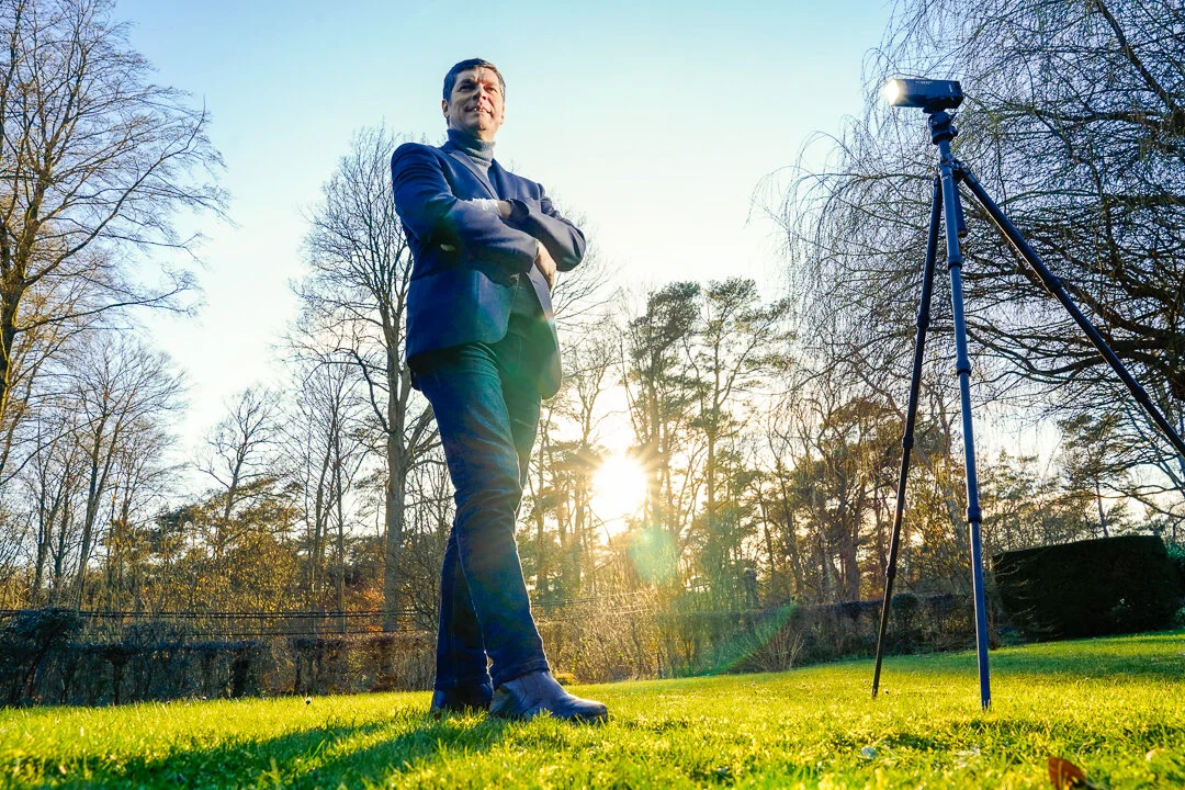Paul Marnef se tient dans un jardin près d’un appareil sur trépied, avec le soleil bas entre les arbres.