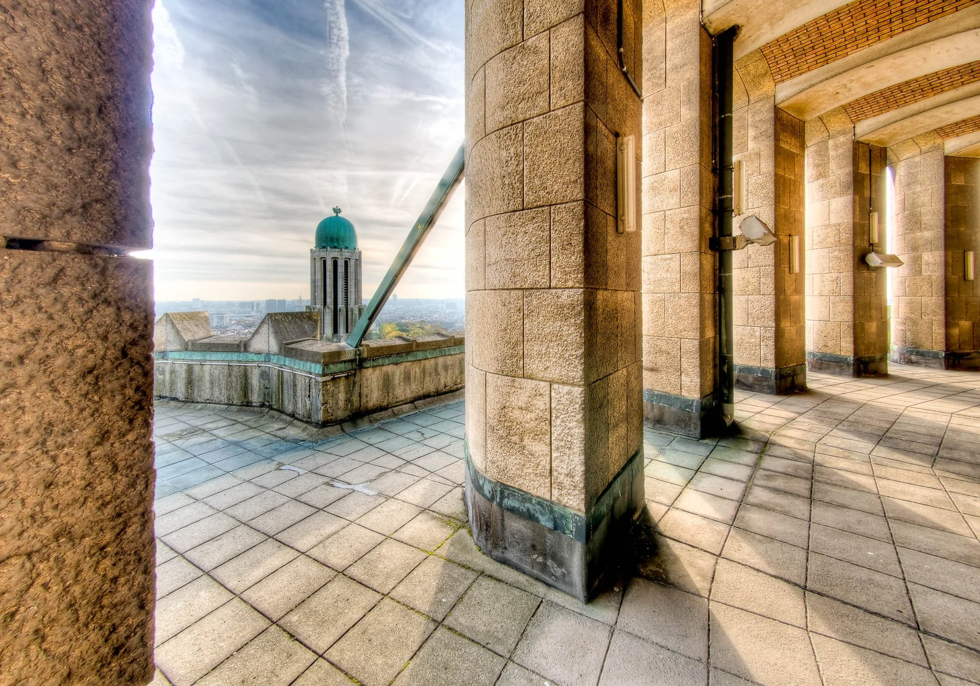 Vue du sommet d'un bâtiment avec une terrasse en pierre, vue sur la ville avec une tour caractéristique avec un dôme vert, sous un ciel nuageux.