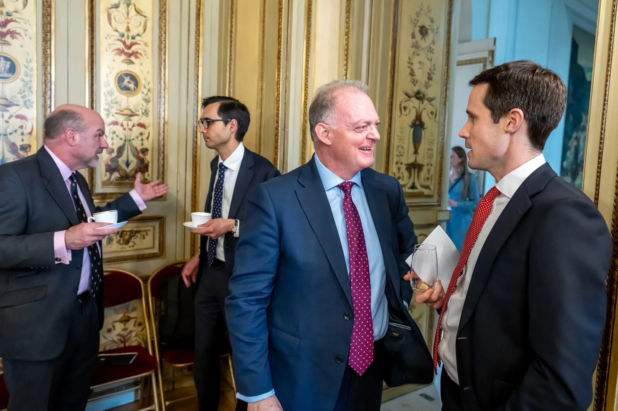 Four men in suits are chatting in an elegant room, with gold decorative details on the walls. Two of the men are holding cups of coffee, and another is holding a glass.