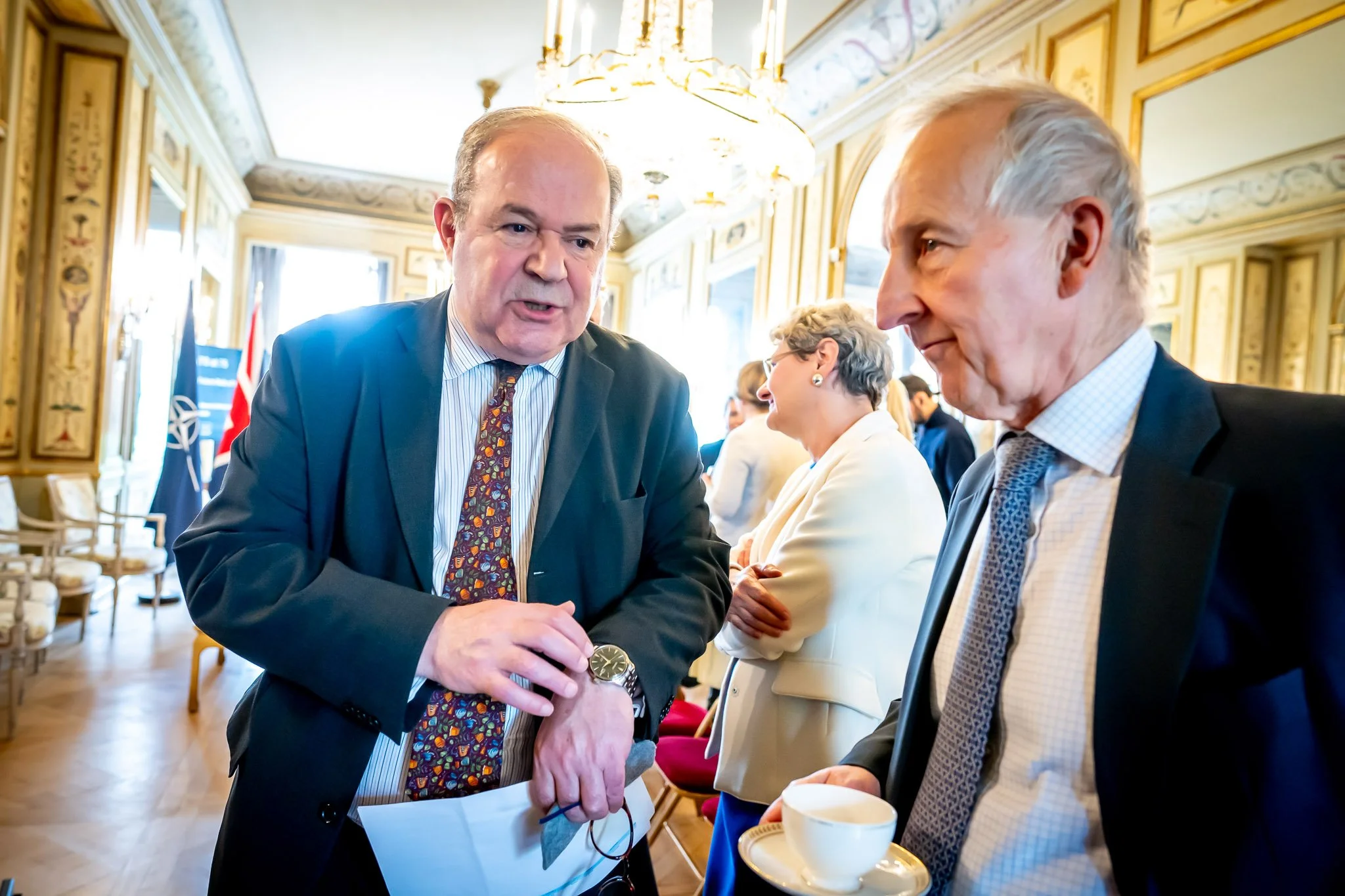 Two men in suits are talking in an elegant room, with other people in the background. One is holding a cup of coffee and the other is looking at his watch. A NATO flag can be seen in the background.