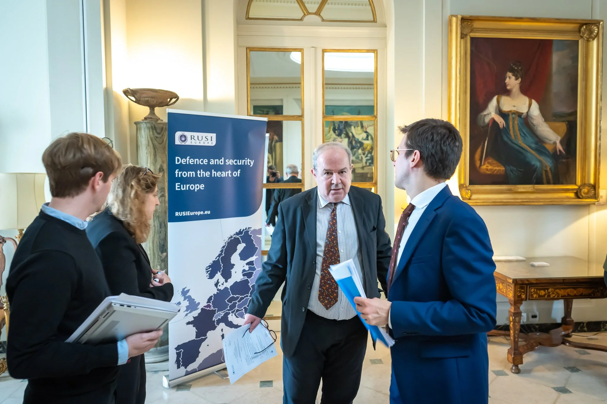 Several people are chatting around a “RUSI Europe” banner bearing the words “Defence and security from the heart of Europe” in a room decorated with paintings.