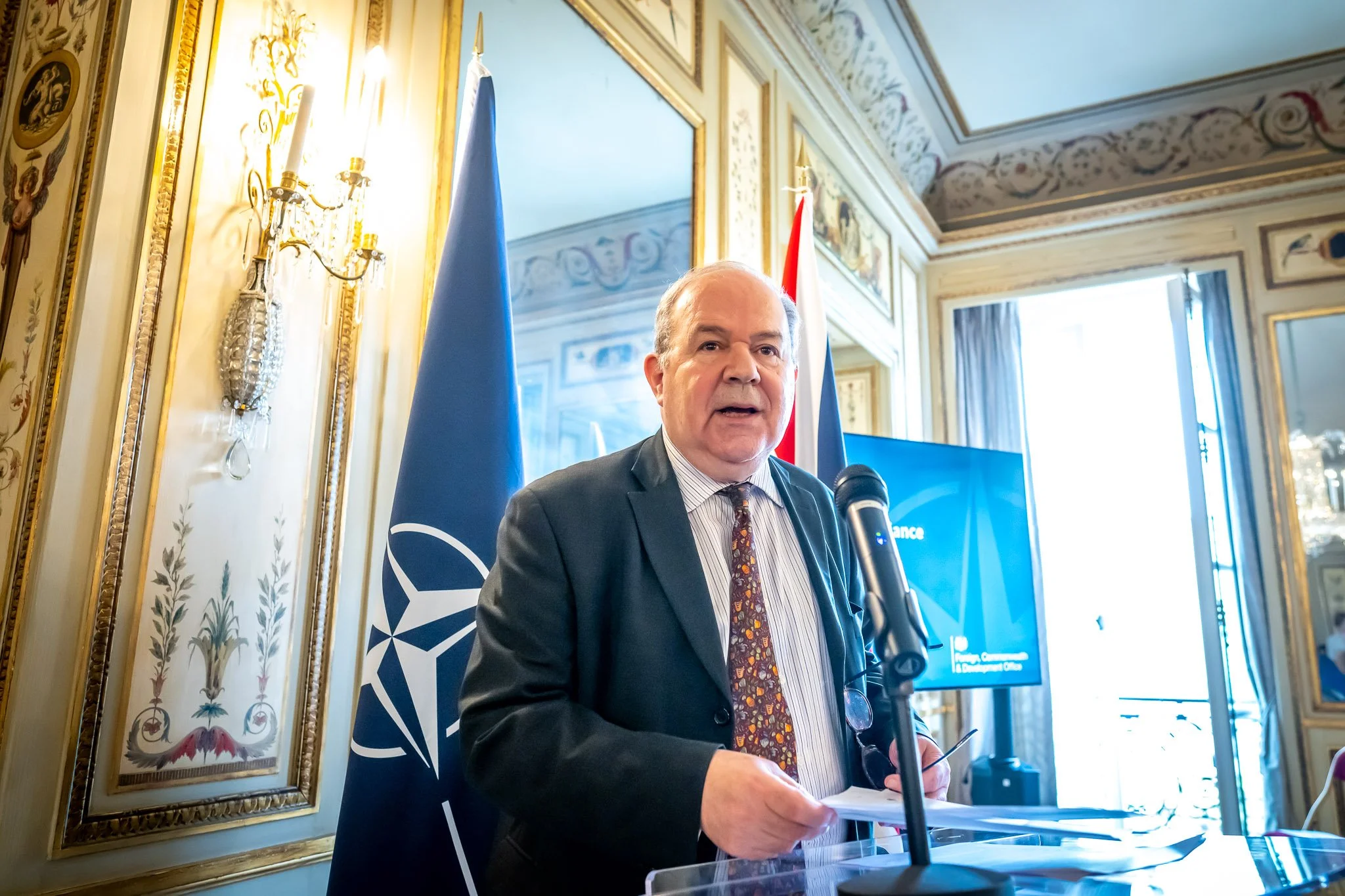 A man in a suit delivering a speech at a microphone, with a NATO flag in the background in a decorated room.