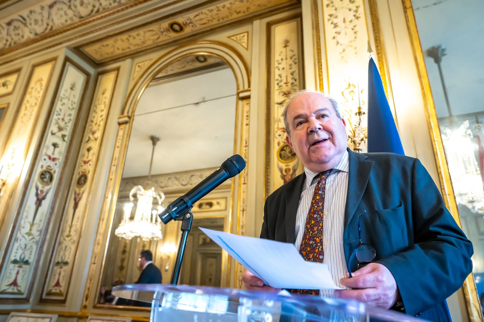 A man speaking into a microphone in a room adorned with mirrors and gilded decorations.