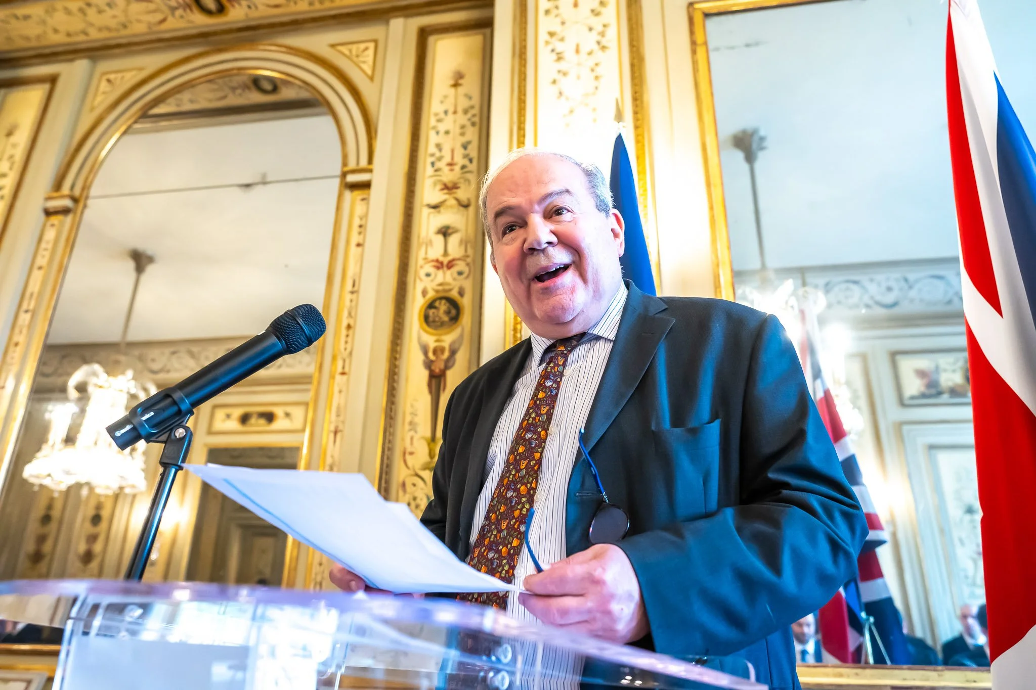 A smiling man holding some papers behind a lectern with a microphone, with flags in the background.
