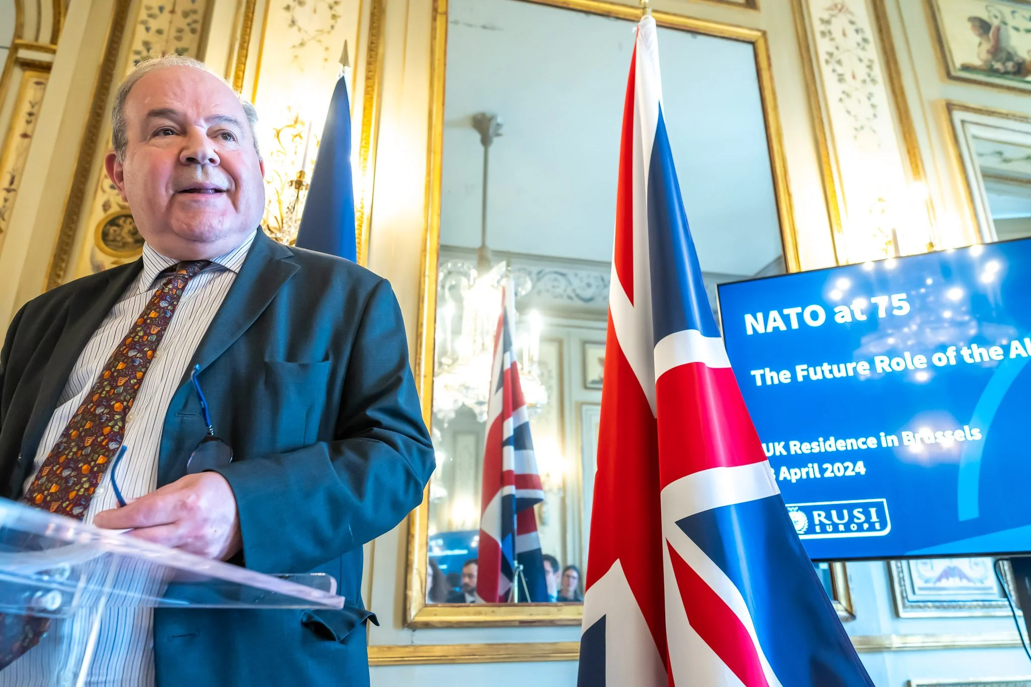 A man in a suit speaking at a NATO event in Brussels, with a British flag and a screen displaying ‘NATO at 75’.