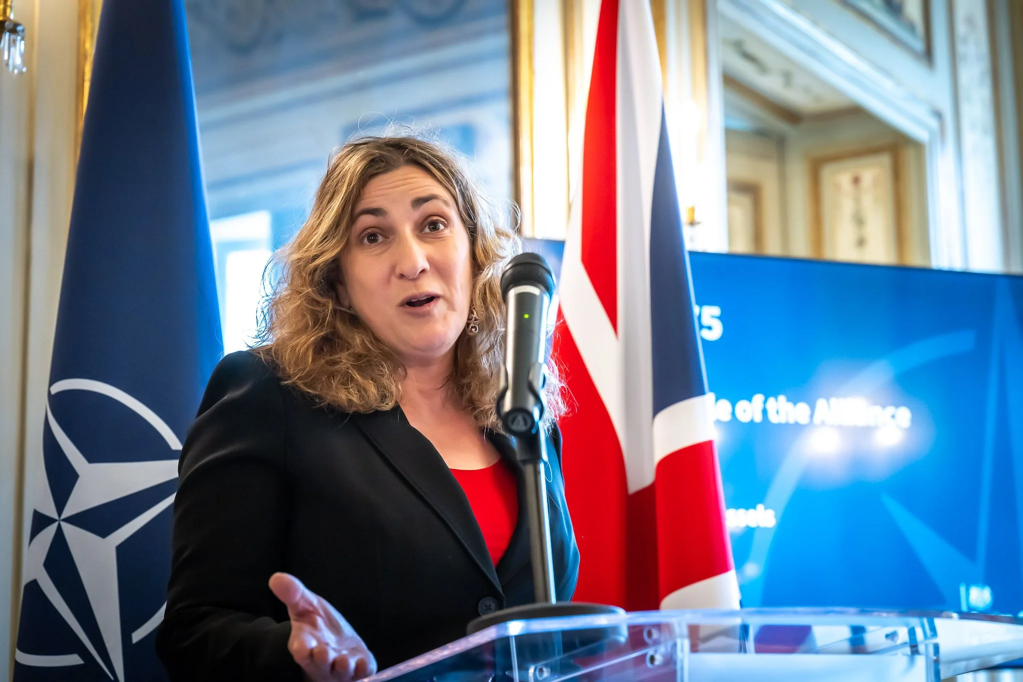 A woman is speaking at a lectern with a microphone; in the background are a British flag and a NATO flag.