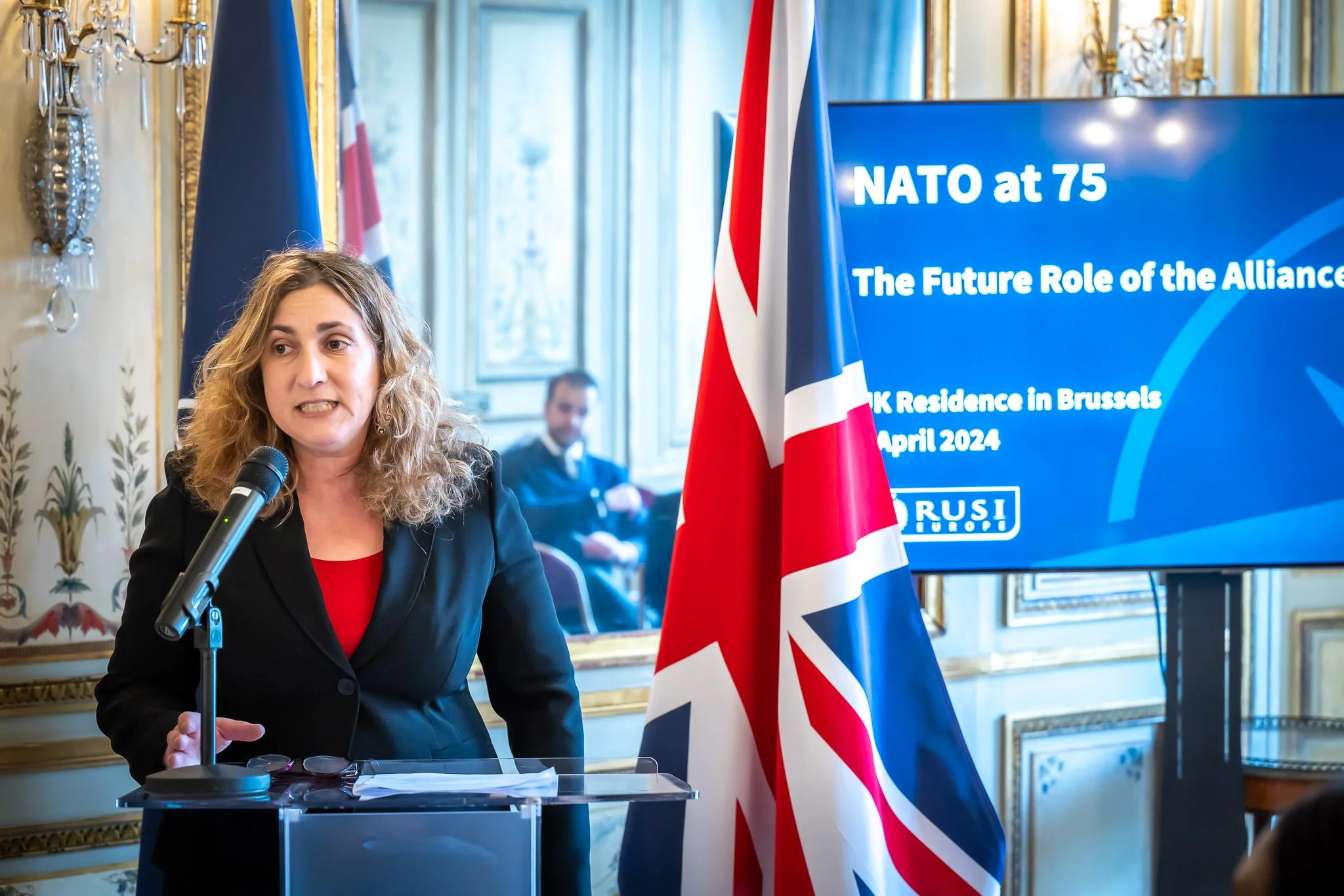 A woman speaking into a microphone in an ornate room, with the British flag in the foreground; a screen displaying ‘NATO at 75 – The Future Role of the Alliance’, the Union Jack visible, and candlesticks in the background.