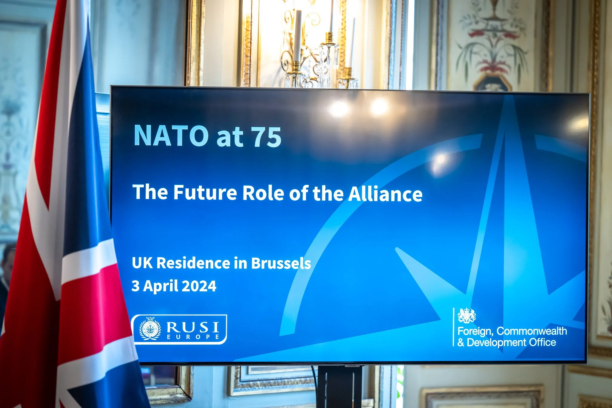 A slide displaying ‘NATO at 75’ and ‘The Future Role of the Alliance’ at a UK event in Brussels, with a British flag in the background, on 3 April 2024.