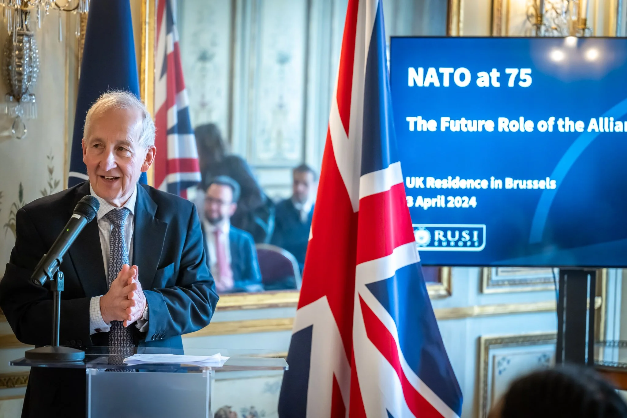 Un homme en costume parle à un micro lors d'un événement, avec un écran derrière lui affichant « NATO at 75 » et un drapeau britannique à côté.