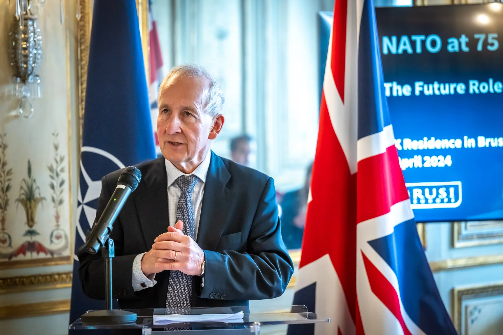 A speaker in a suit is speaking into a microphone, with NATO and UK flags in the background, at an event marking NATO’s 75th anniversary.