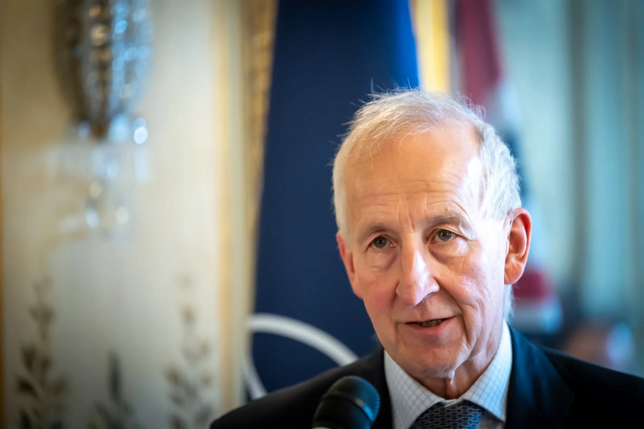 Portrait of an older man in a dark suit and tie at an indoor conference.