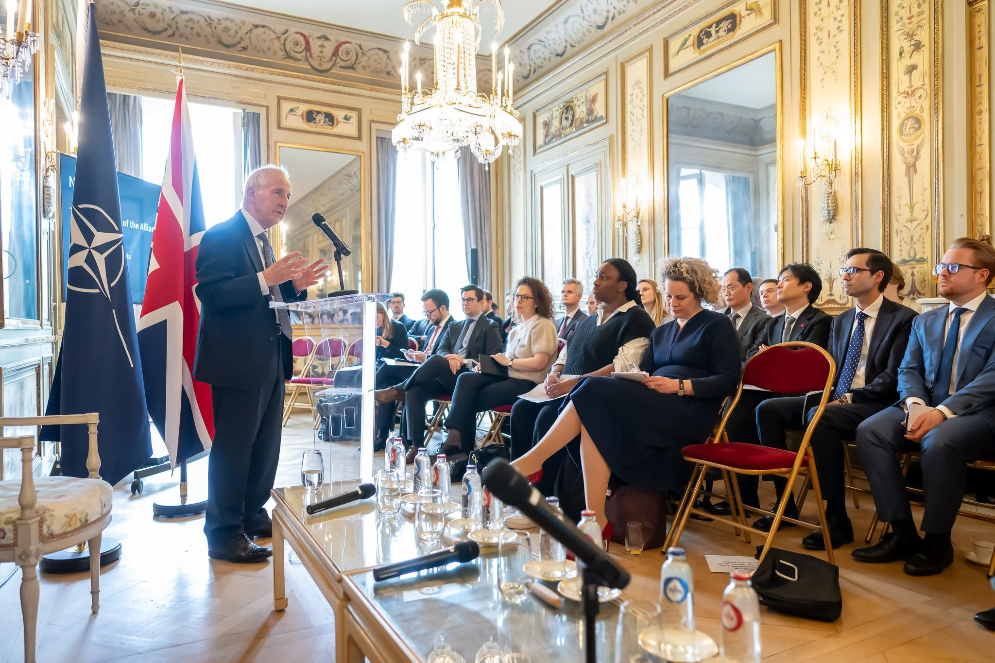 A man is giving a speech to an audience seated in an elegant hall with mirrors and candlesticks. NATO and UK flags can be seen behind him.