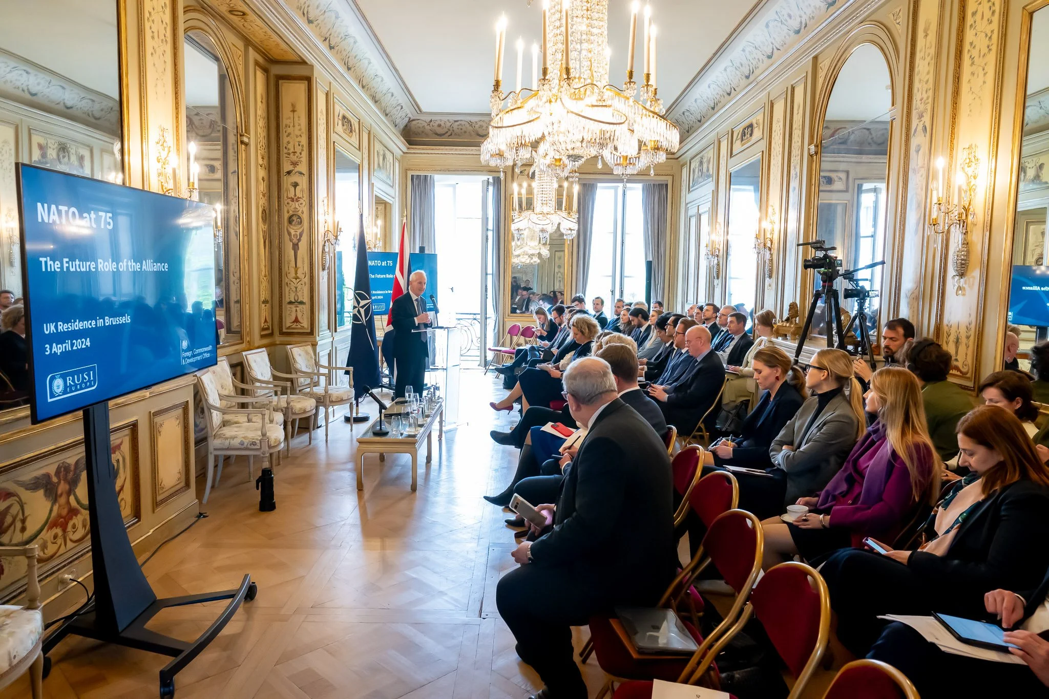 A conference room with participants seated, listening to a presentation on NATO to mark its 75th anniversary, in Brussels. A screen displays details of the event. The room is lavishly decorated with mirrors and a large chandelier.