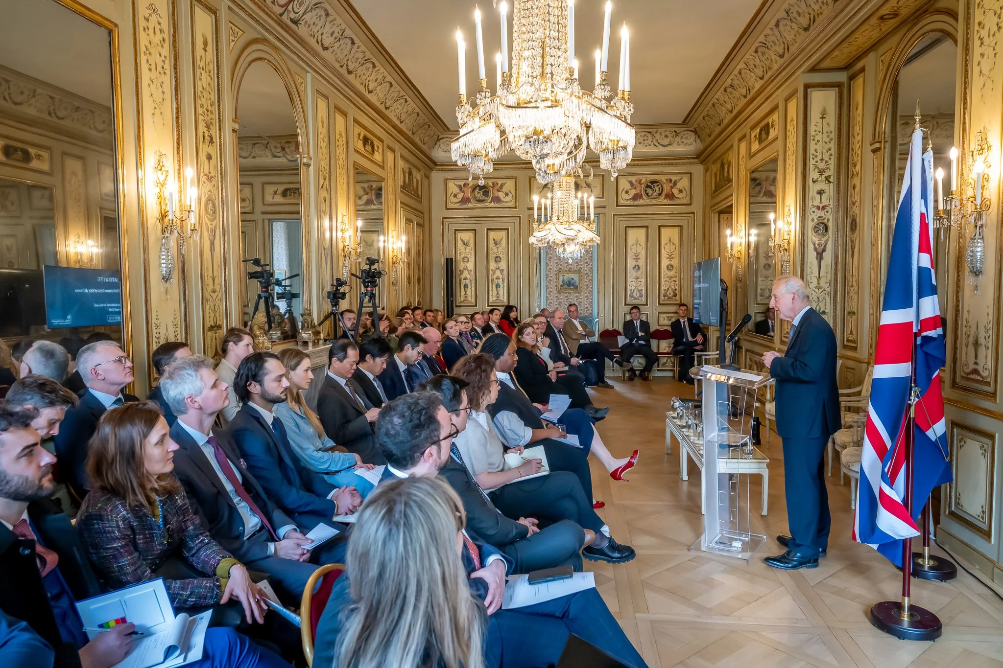 A man is giving a speech in a room decorated with candlesticks and mirrors, in front of a seated audience. British flags can be seen.