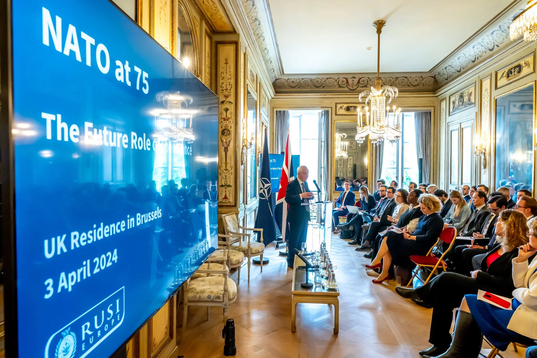 A conference on NATO’s 75th anniversary at the UK Residence in Brussels, with a speaker addressing a seated audience. A large slide on the screen displays the date 3 April 2024. An elegant interior setting with chandeliers and mirrors.