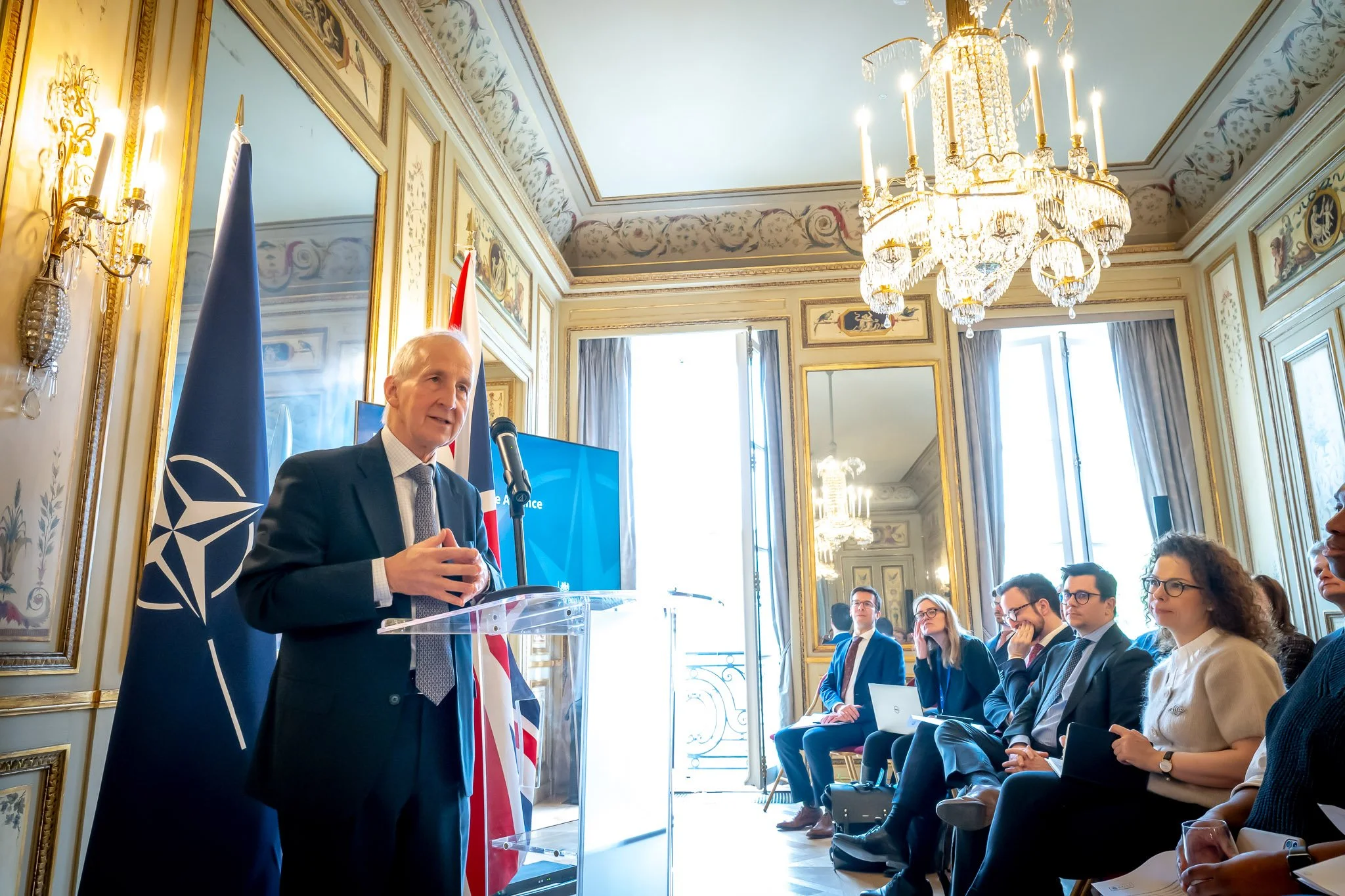 A man in a suit is speaking into a microphone at a conference, with NATO and UK flags in the background. People are seated and listening attentively in an elegant room with chandeliers and mirrors.