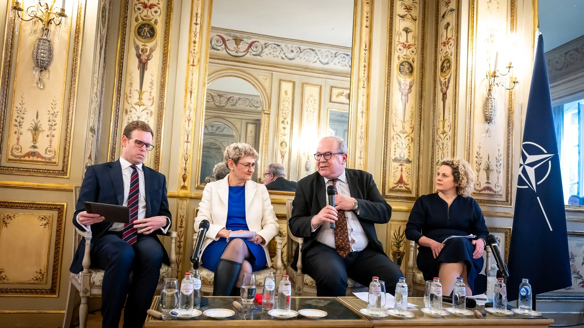 Four people sitting in a decorated room in front of a NATO flag, speaking at a conference, with microphones and bottles of water on a table.