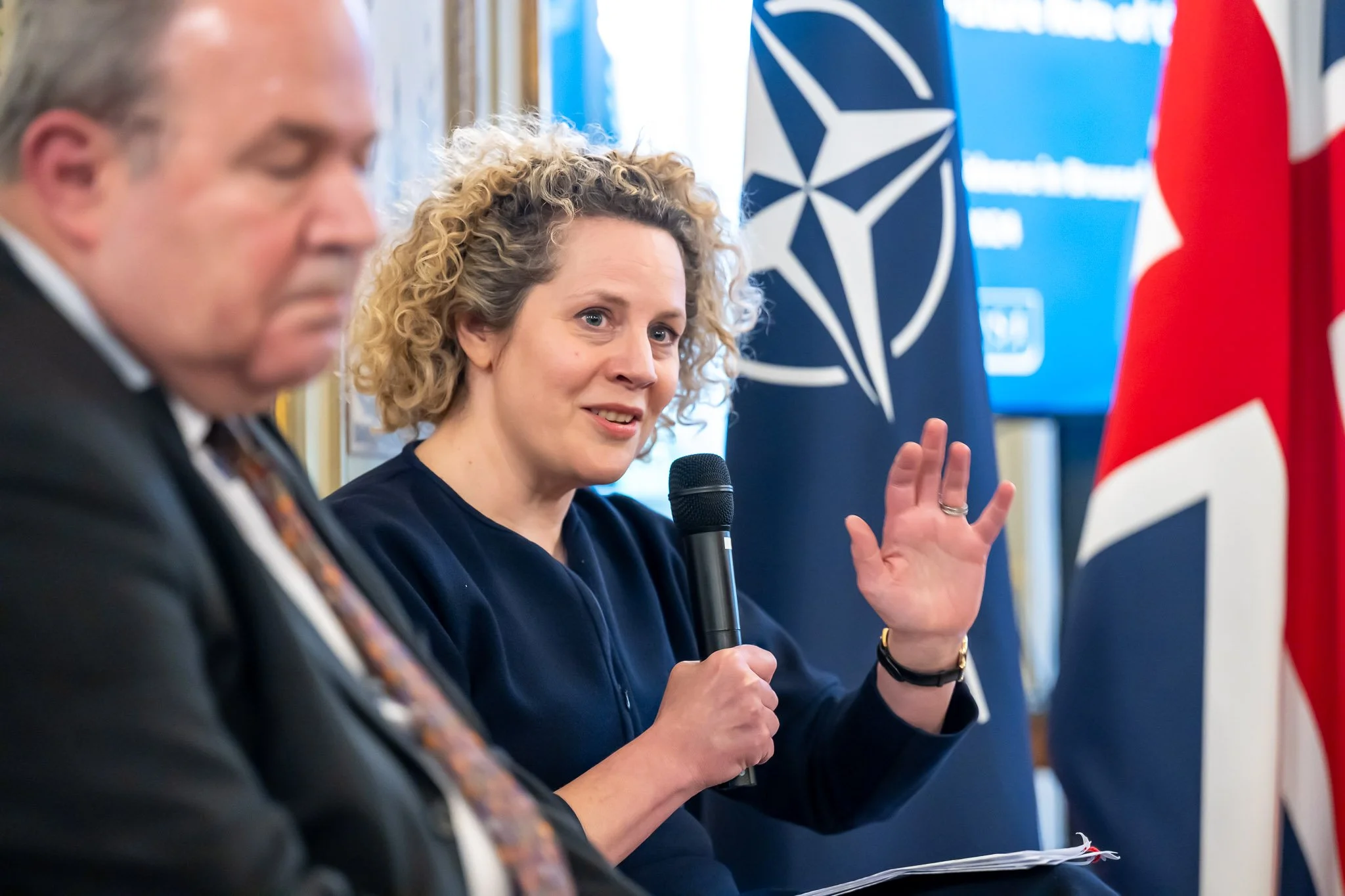 A woman with curly hair gestures while speaking into a microphone during a panel discussion.