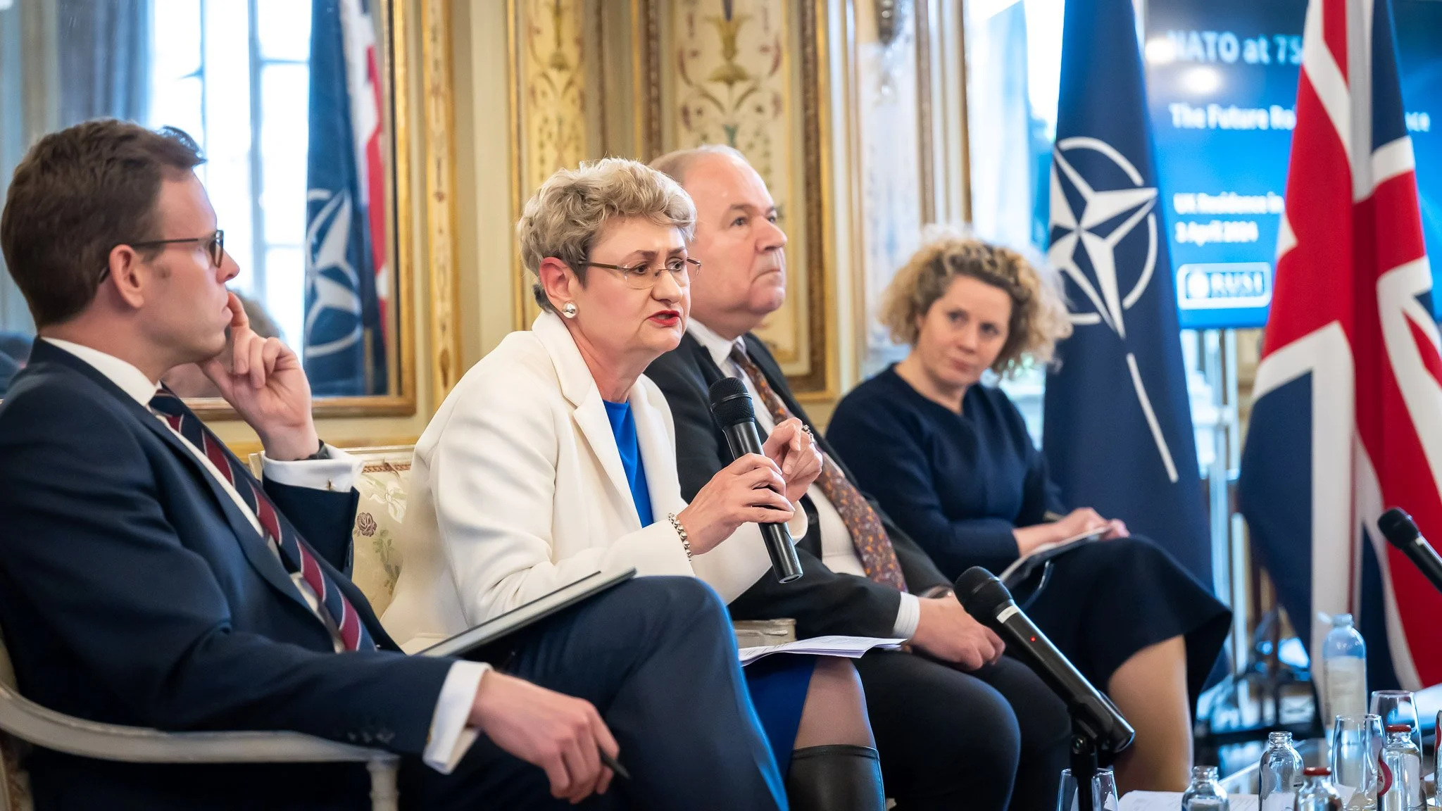People seated at a conference, with a woman holding a microphone. In the background, NATO and UK flags can be seen. A poster reads ‘NATO at 75’ and ‘The Future of...’. The participants appear to be in a