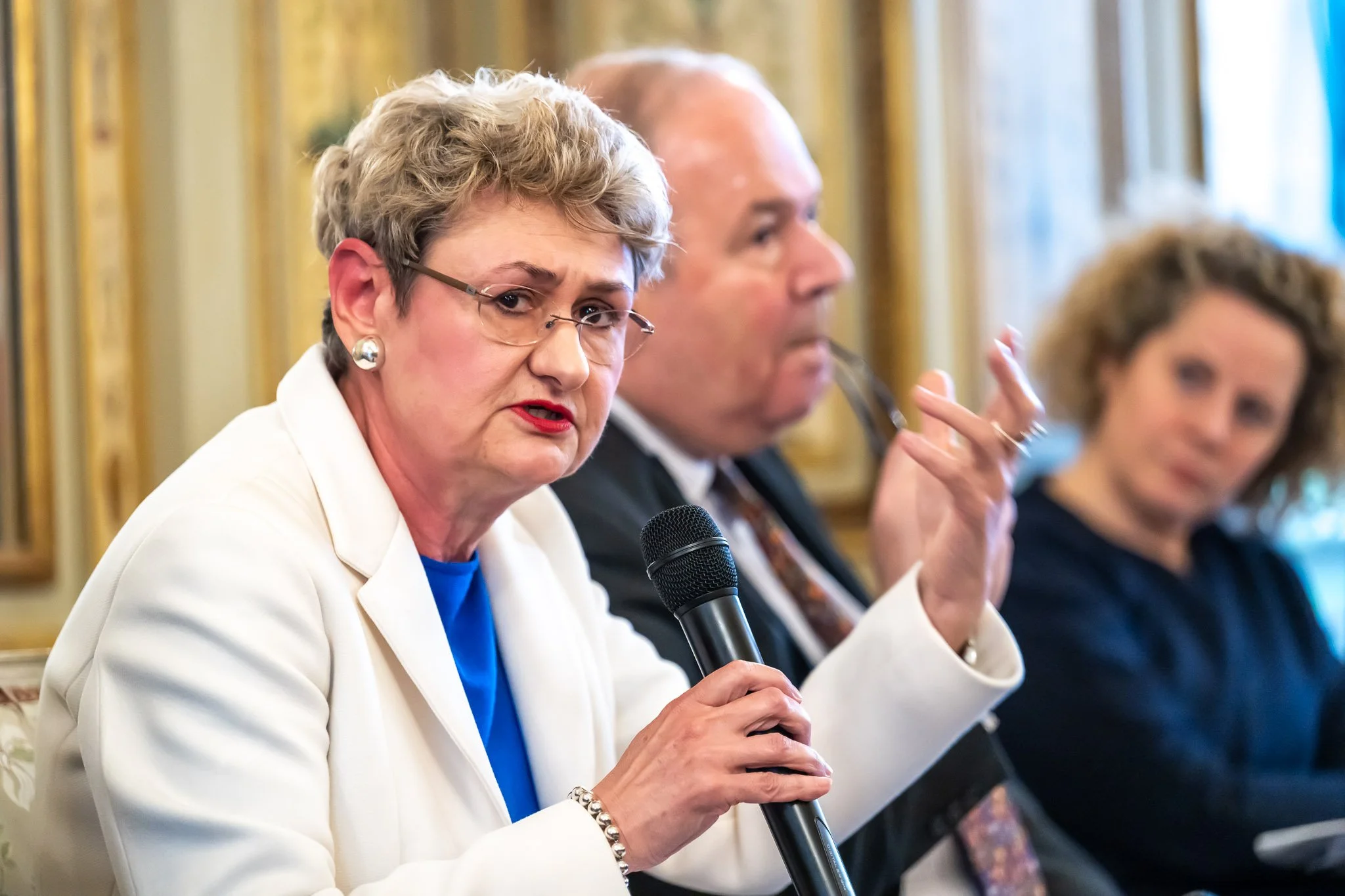 A woman holding a microphone speaking at an event, with two other people blurred in the background.