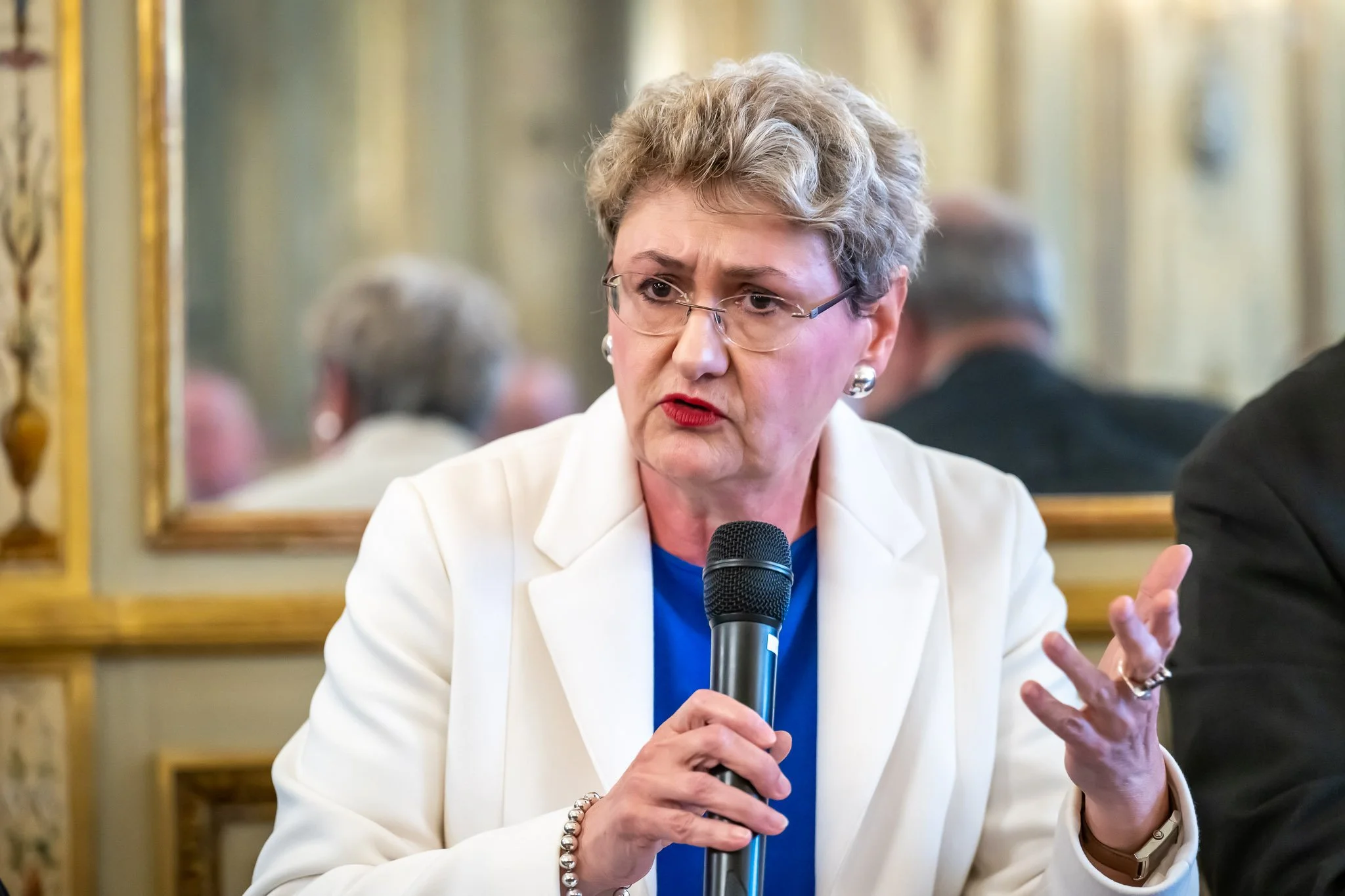 A woman holding a microphone, wearing a white suit, speaking at an event.