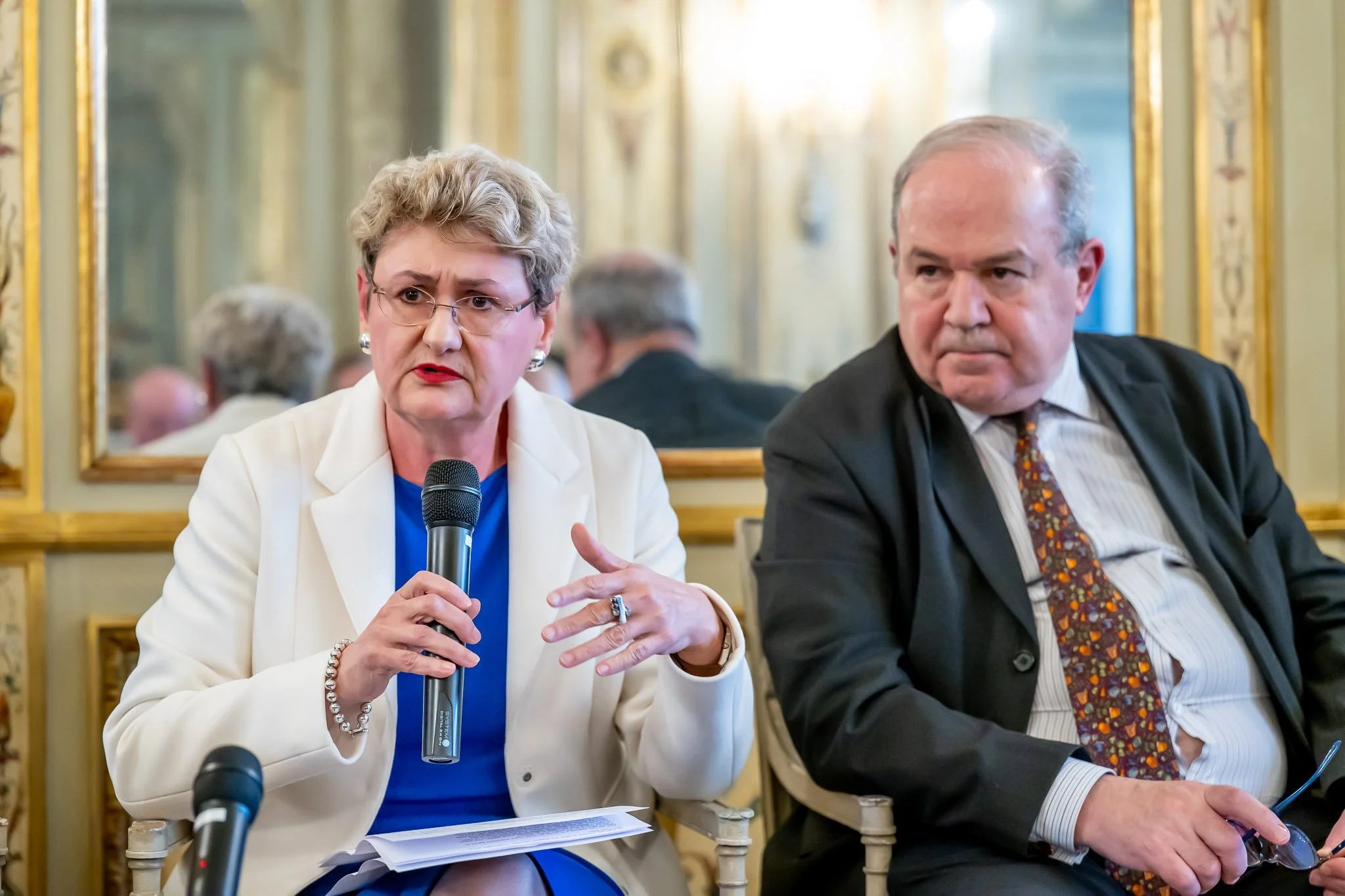 Two people are sitting and talking. The woman on the left is holding a microphone and is wearing a white suit. The man on the right is wearing a dark suit with a tie. Behind them is a wall with a mirror and elegant decorations.
