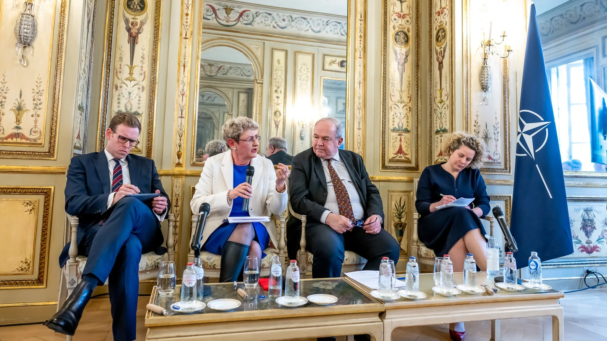 Four people are seated in a decorated room, with a NATO flag on the right. Two women are talking whilst two men listen, surrounded by bottles of water and microphones.