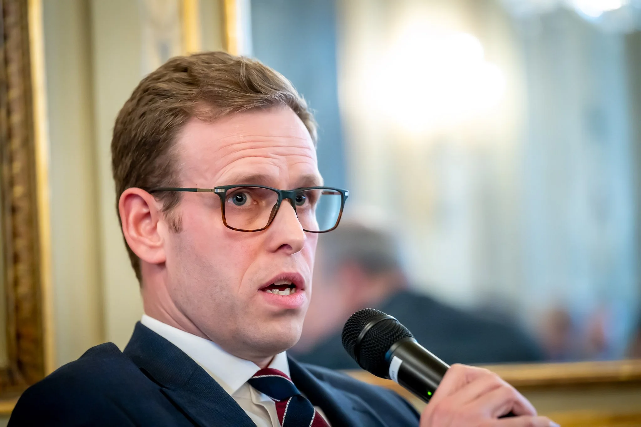 A man wearing glasses, holding a microphone, probably speaking at an event.