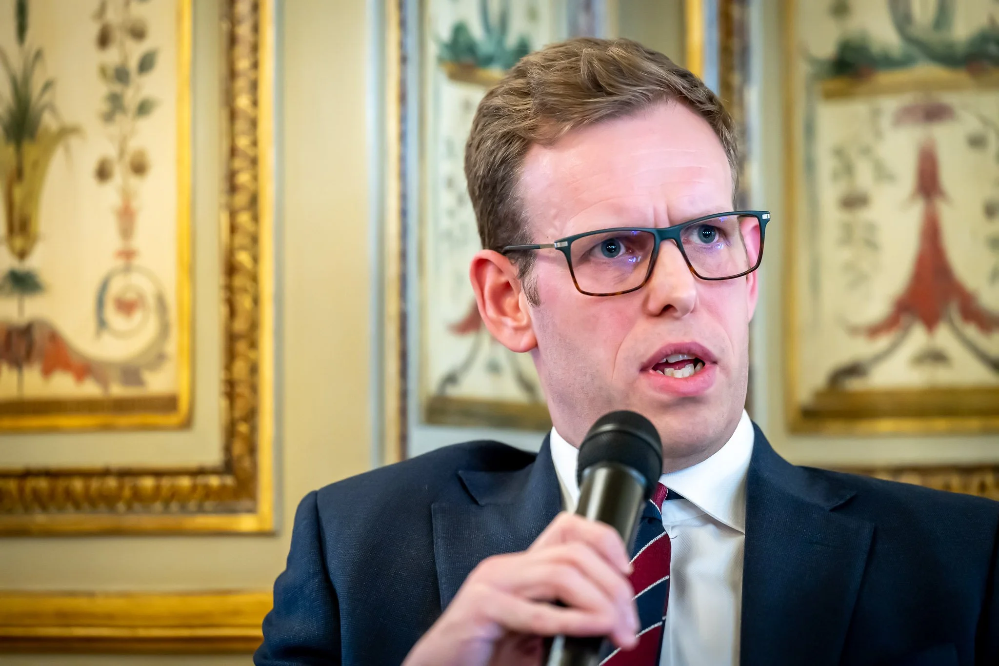 A man in a suit is speaking into a microphone, against a backdrop of an elegant interior and gilded frames.