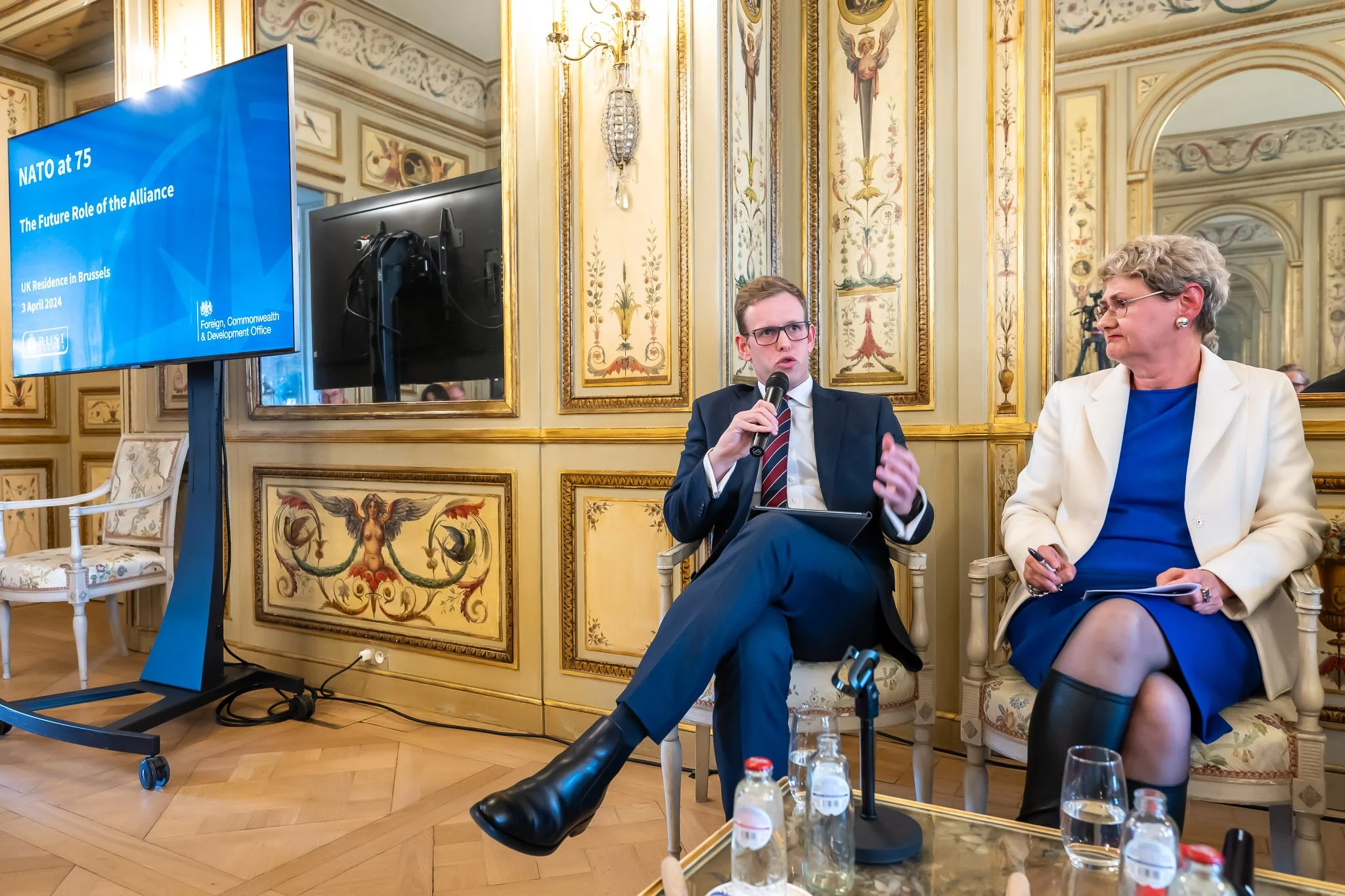Two people in conversation, vintage interior décor, elaborate setting, a sign reading ‘NATO at 75: The Future Role of the Alliance’.