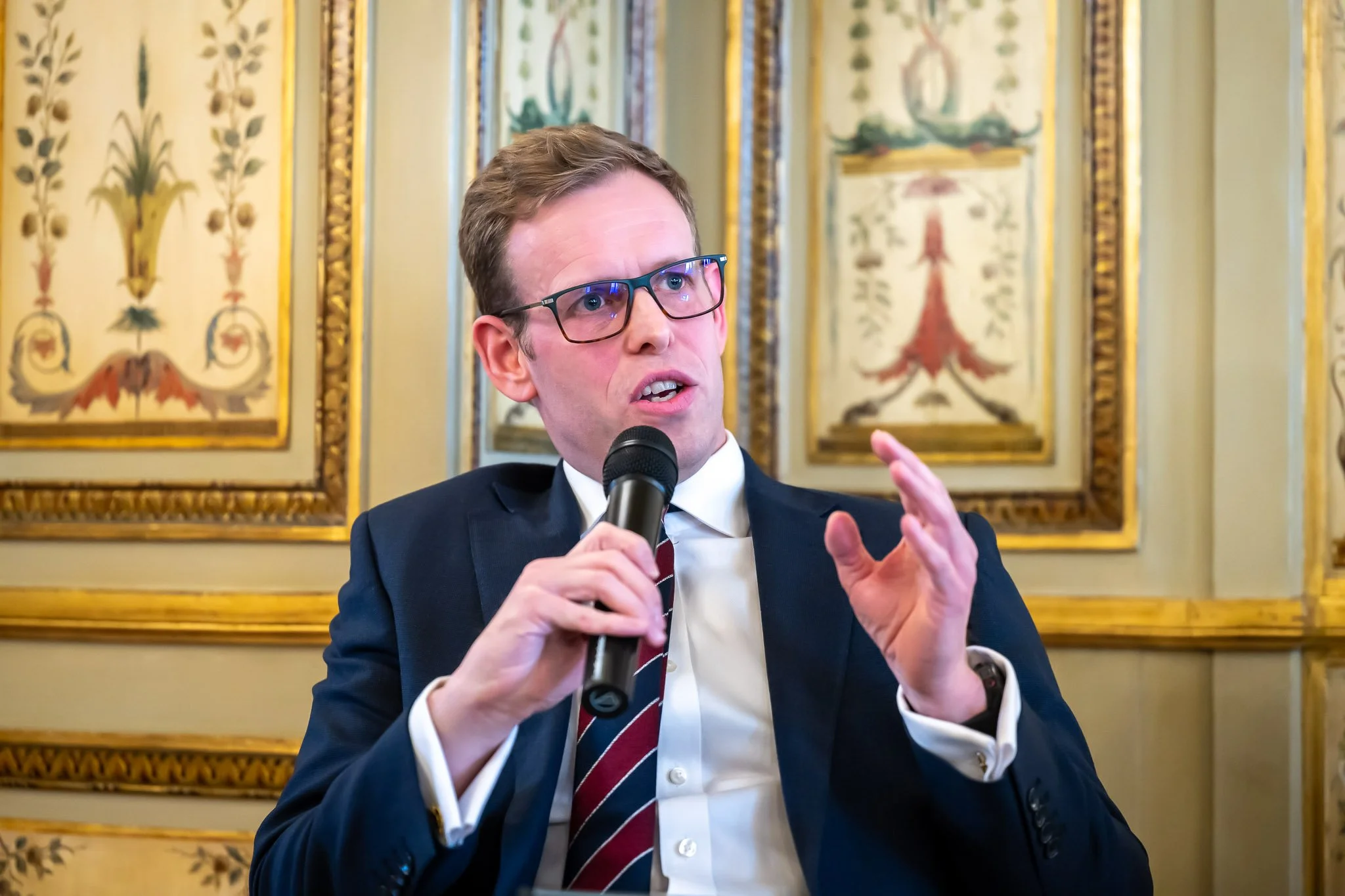 A man in a suit is speaking into a microphone, gesturing with his hand, with an ornate wall decoration in the background.