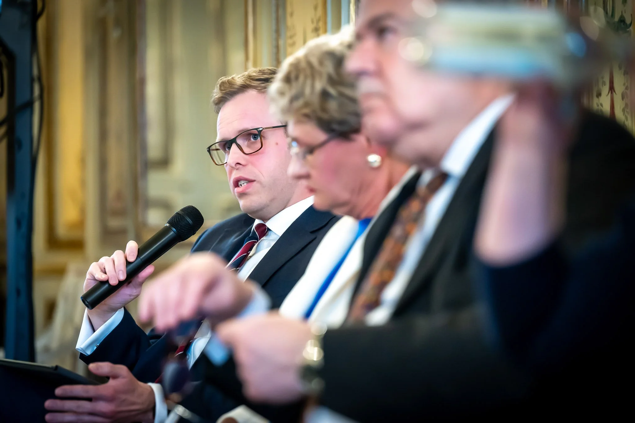 Three people in suits chatting at an event; a man holding a microphone.