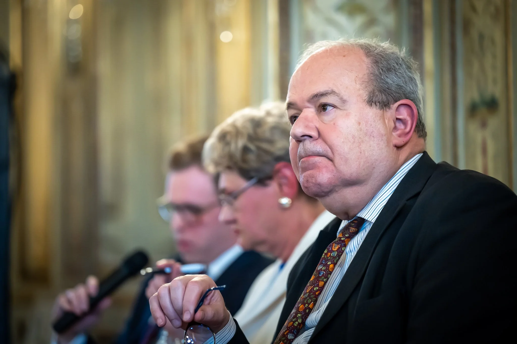 Three people sitting in suits at an official event; one person is holding a pair of glasses.