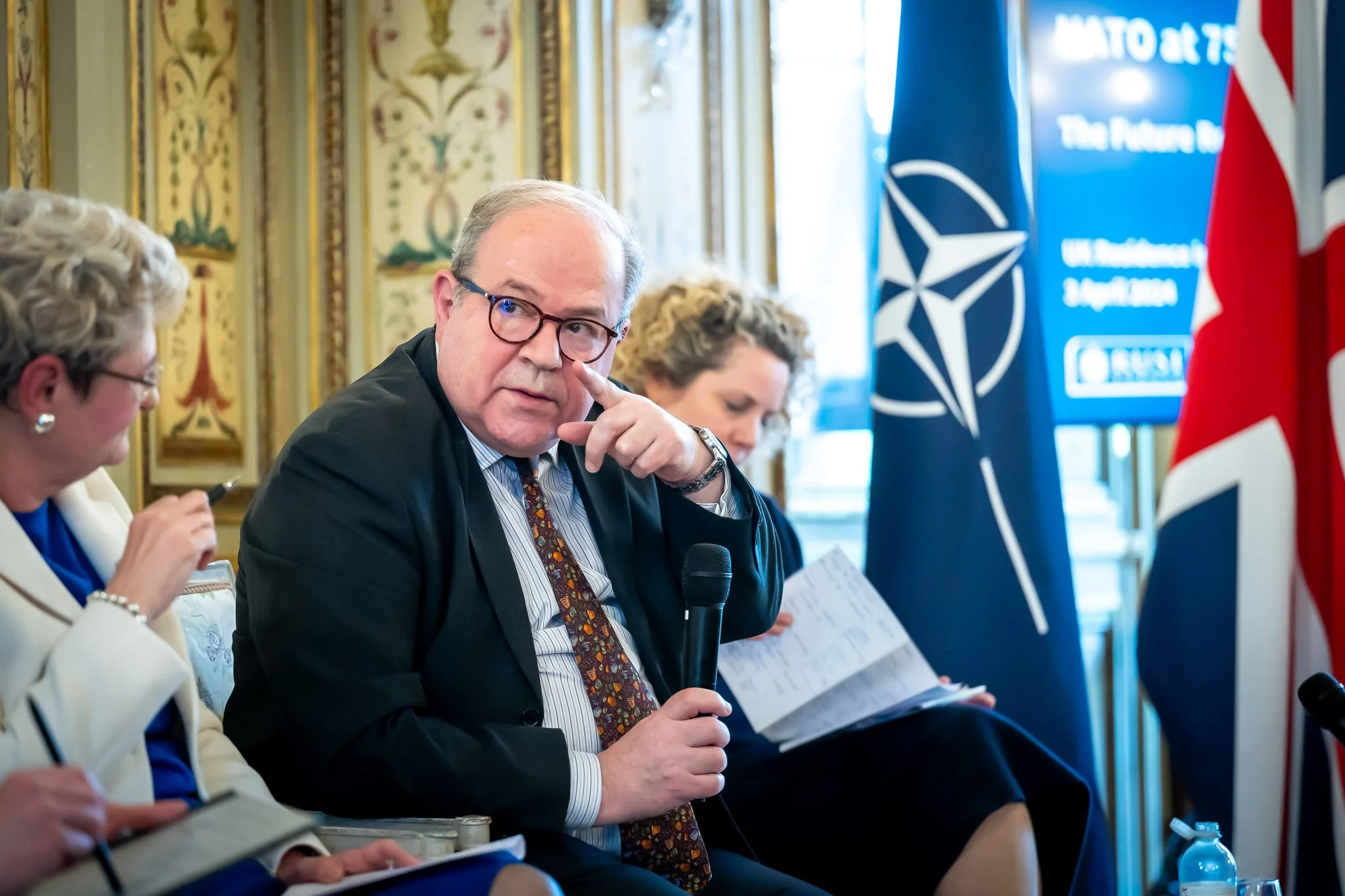 A man in a suit and tie, holding a microphone, speaking at a NATO event with a British flag in the background. Two other people are sitting next to him.