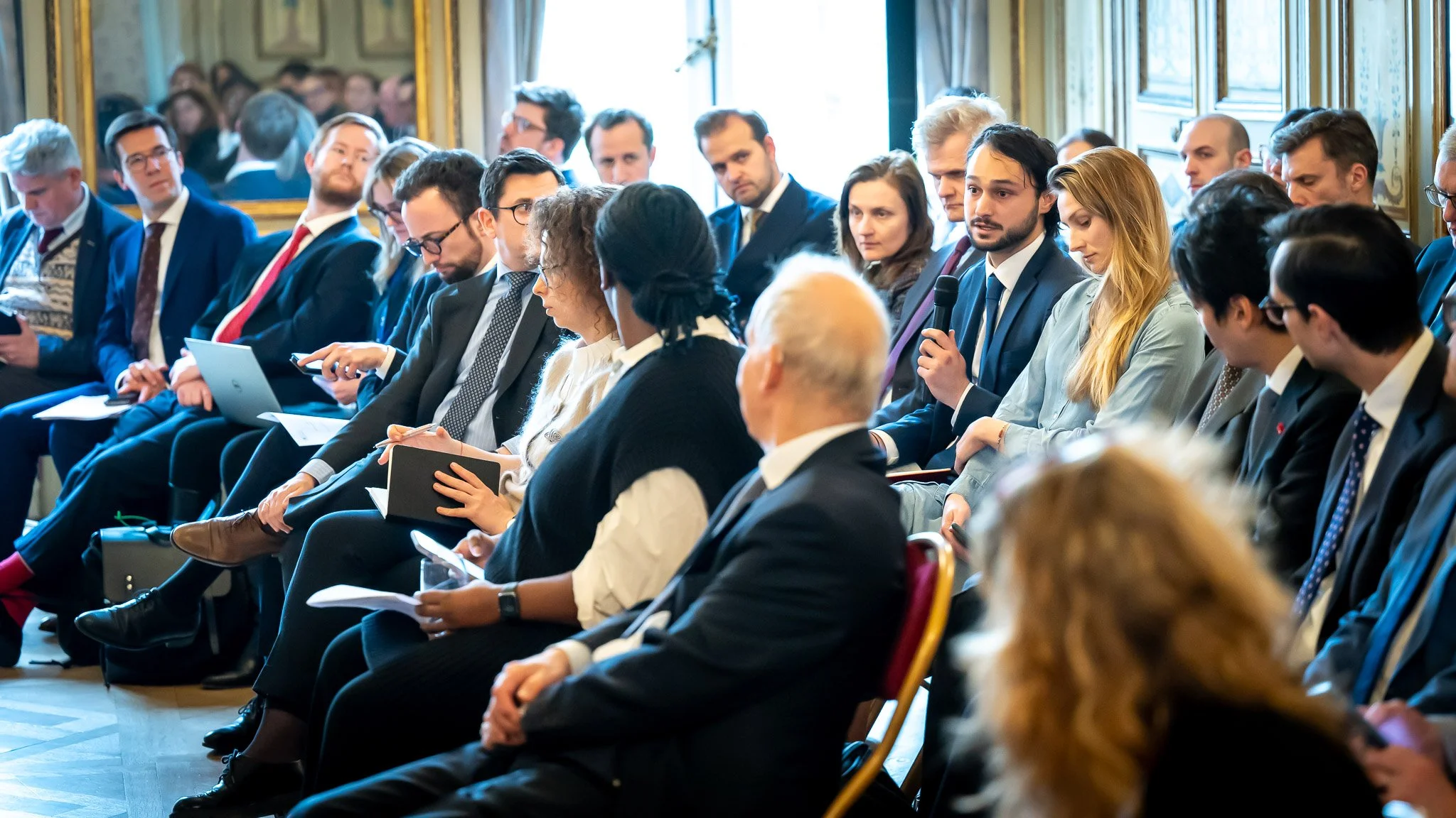 People in suits sitting in a circle at a meeting; a man is speaking into a microphone.