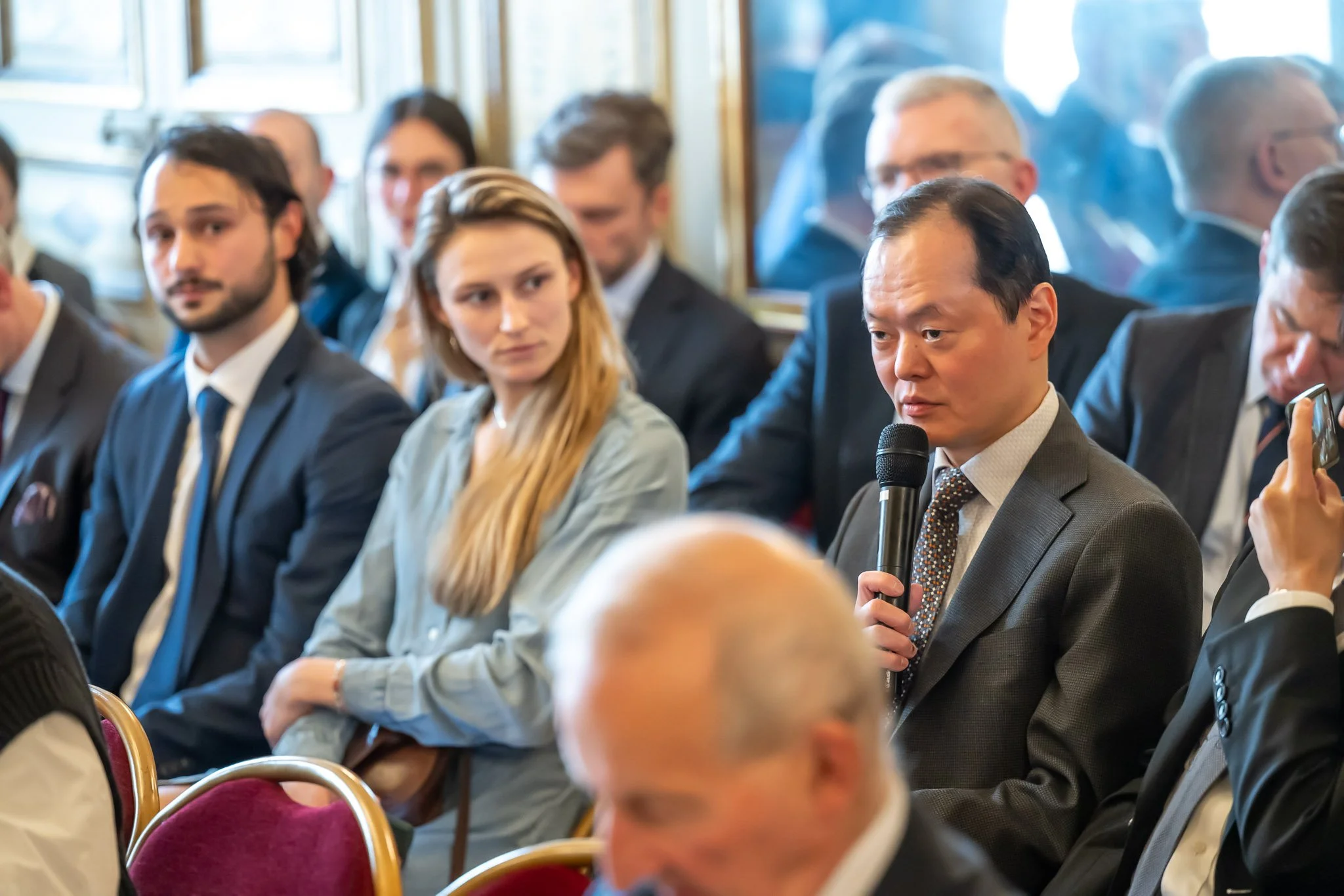 A group of people in suits sitting at a conference, with a man holding a microphone.